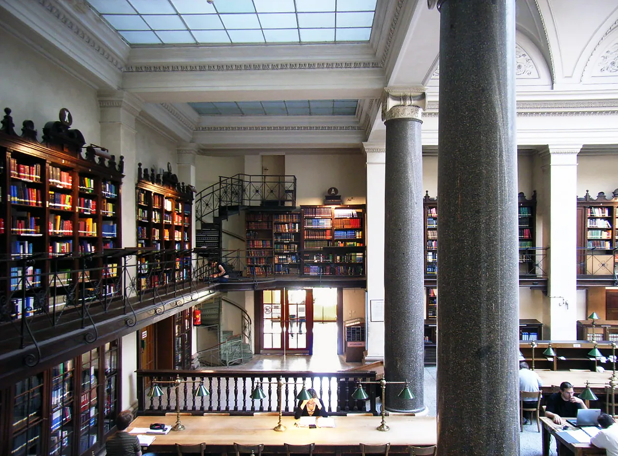 Interior de una gran biblioteca con techos altos, grandes columnas y estanterías llenas de libros. Personas estudiando en escritorios.