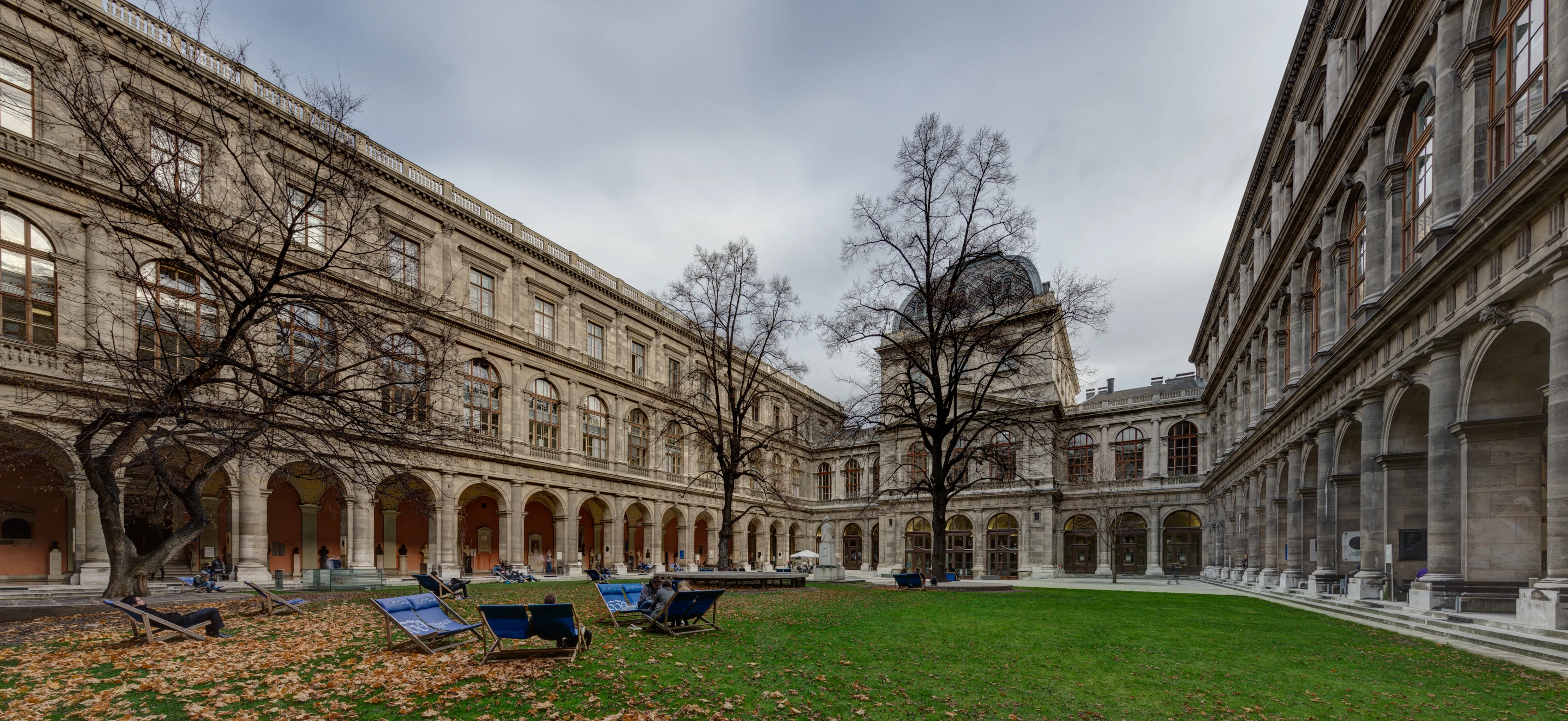 Edificios clásicos con arcadas que rodean un patio ajardinado con árboles desnudos y bancos en la Universidad de Viena
