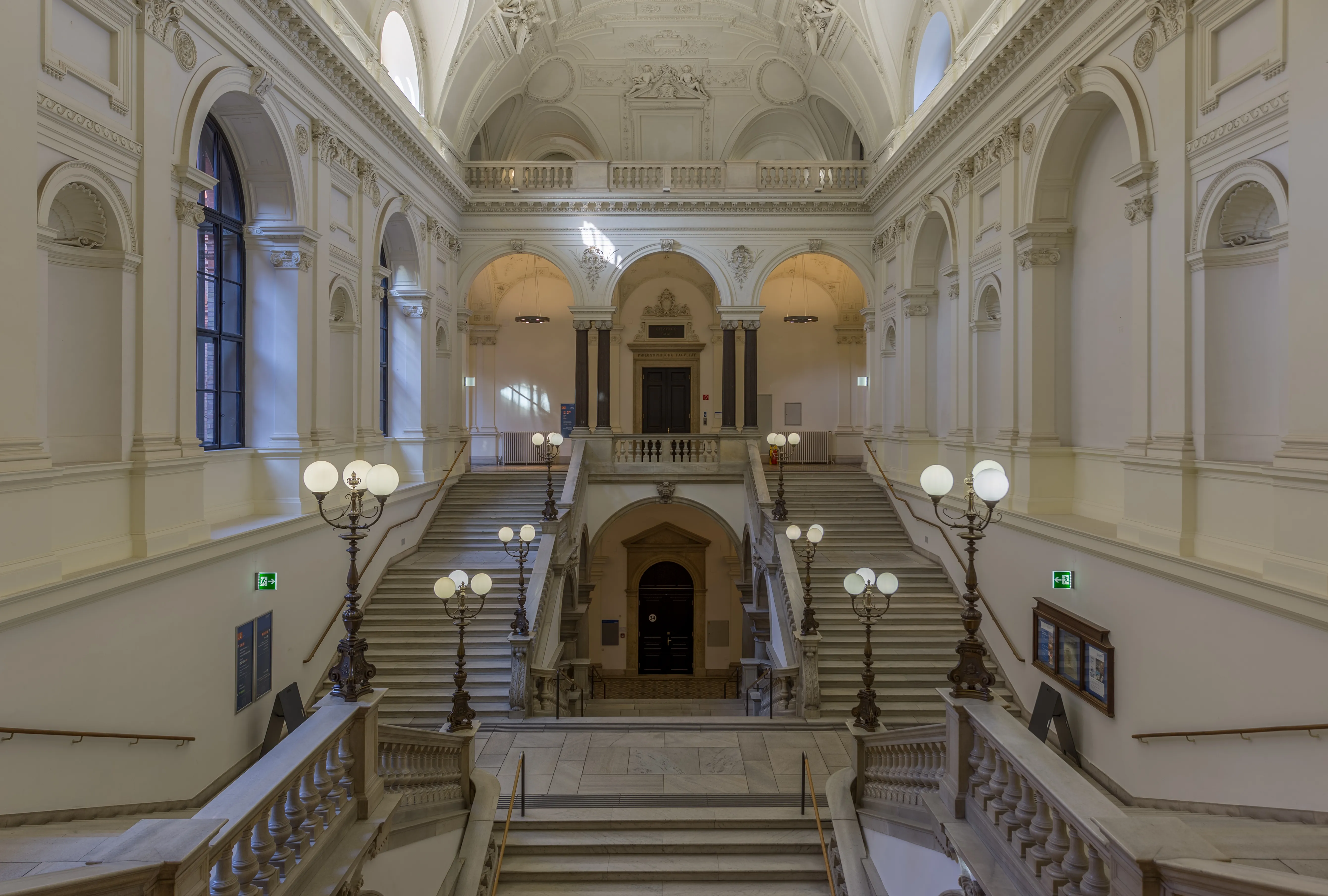 Interior de la Universidad de Viena con una gran escalera central, puertas arqueadas y lámparas ornamentadas