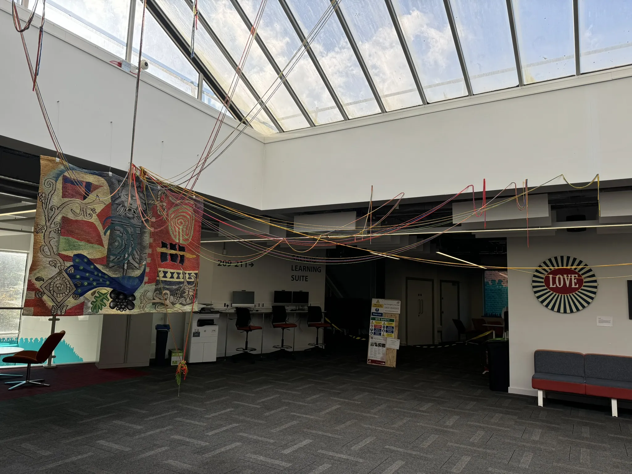Espacio interior del atrio con techo de claraboya de cristal, un gran tapiz textil colgante a la izquierda, cuerdas de colores extendidas por toda la sala, estaciones de trabajo con ordenadores al fondo y un cartel circular de LOVE en la pared de la derecha.