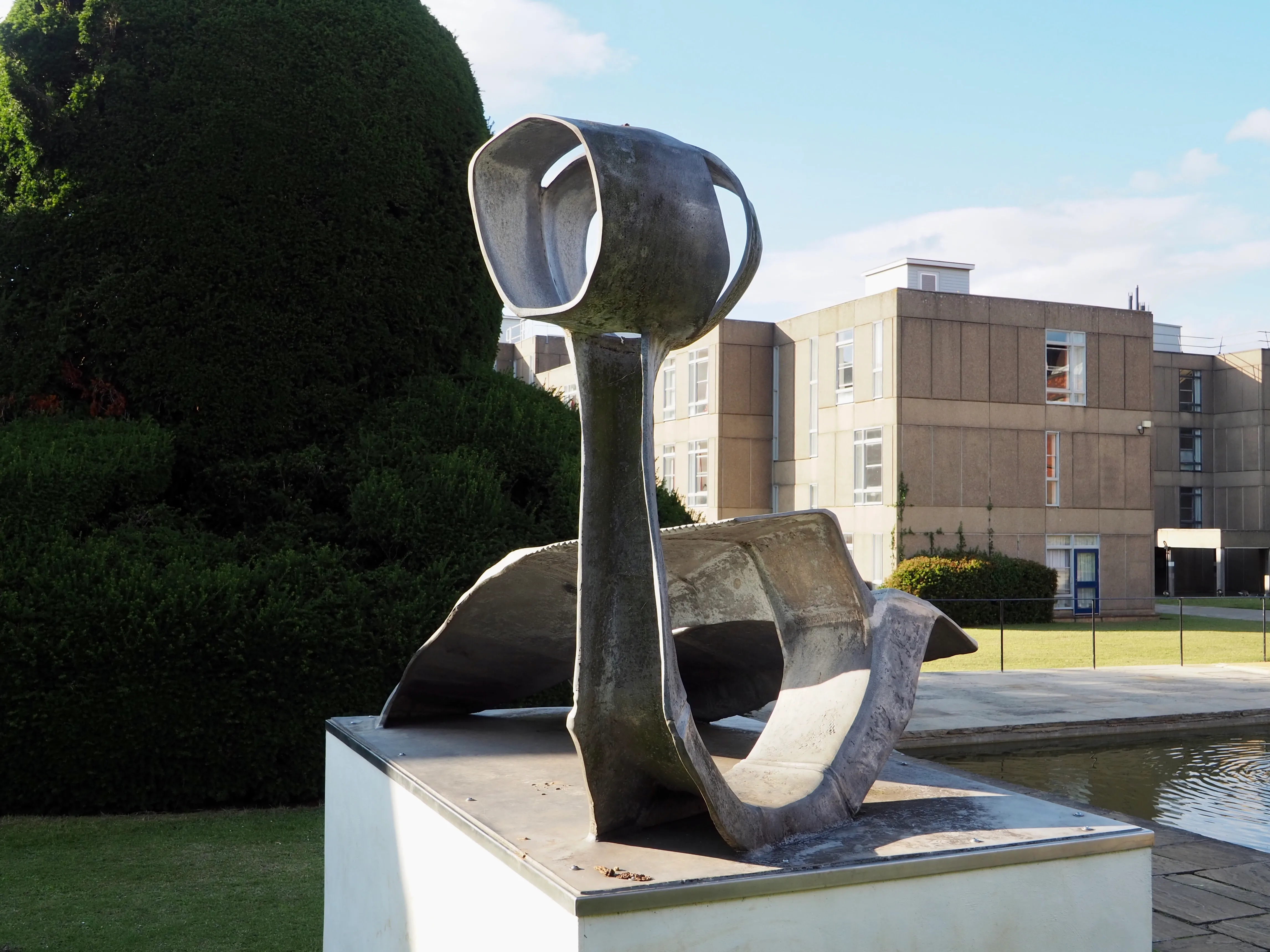Escultura de bronce abstracta sobre un pedestal blanco al aire libre, con una forma alta en forma de copa que surge de una base curva, con un edificio moderno y un seto verde al fondo.
