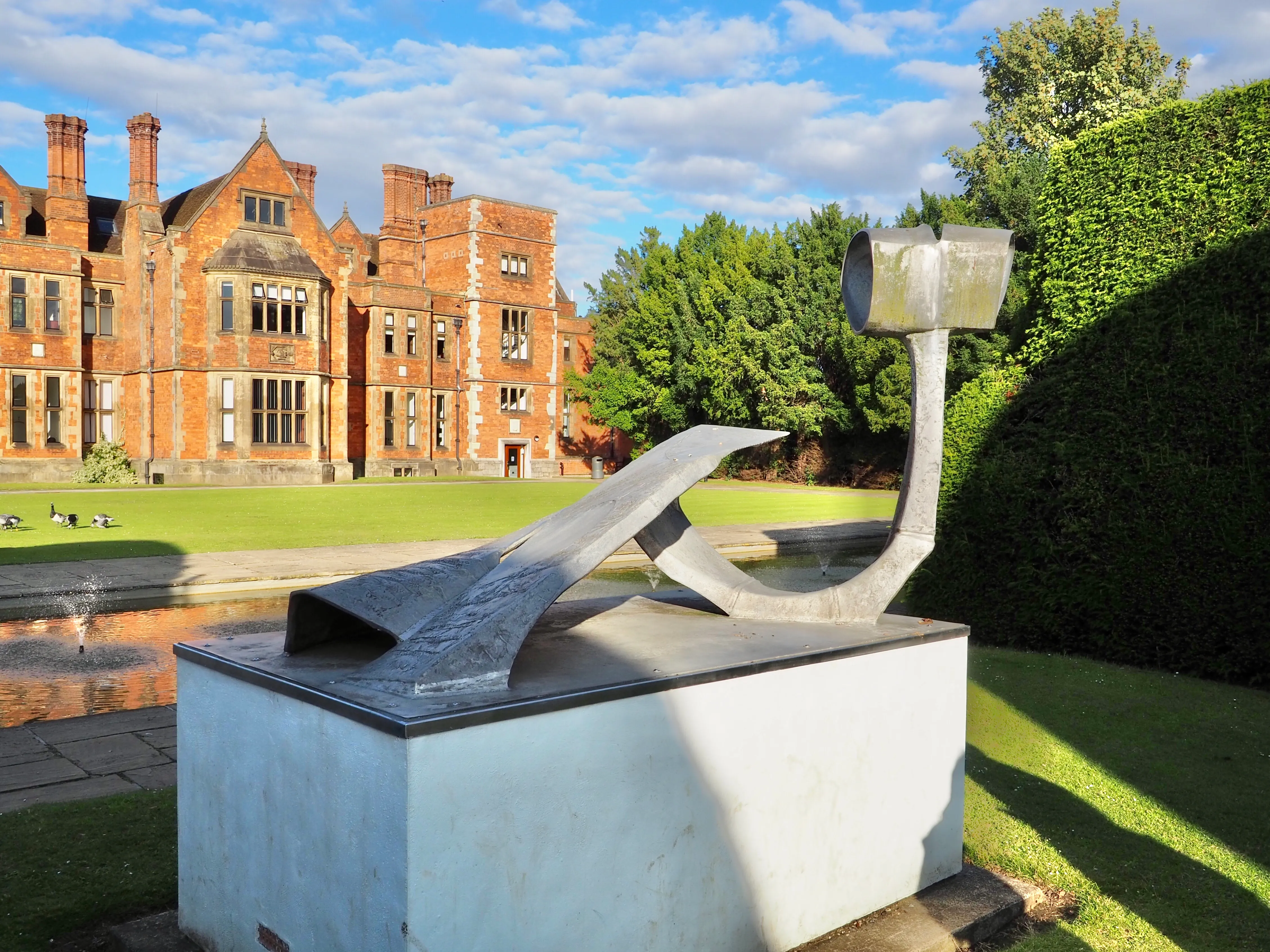 Una escultura abstracta de bronce sobre un pedestal de hormigón situado en el césped de un campus, con un gran edificio victoriano de ladrillo rojo y un estanque visibles al fondo.