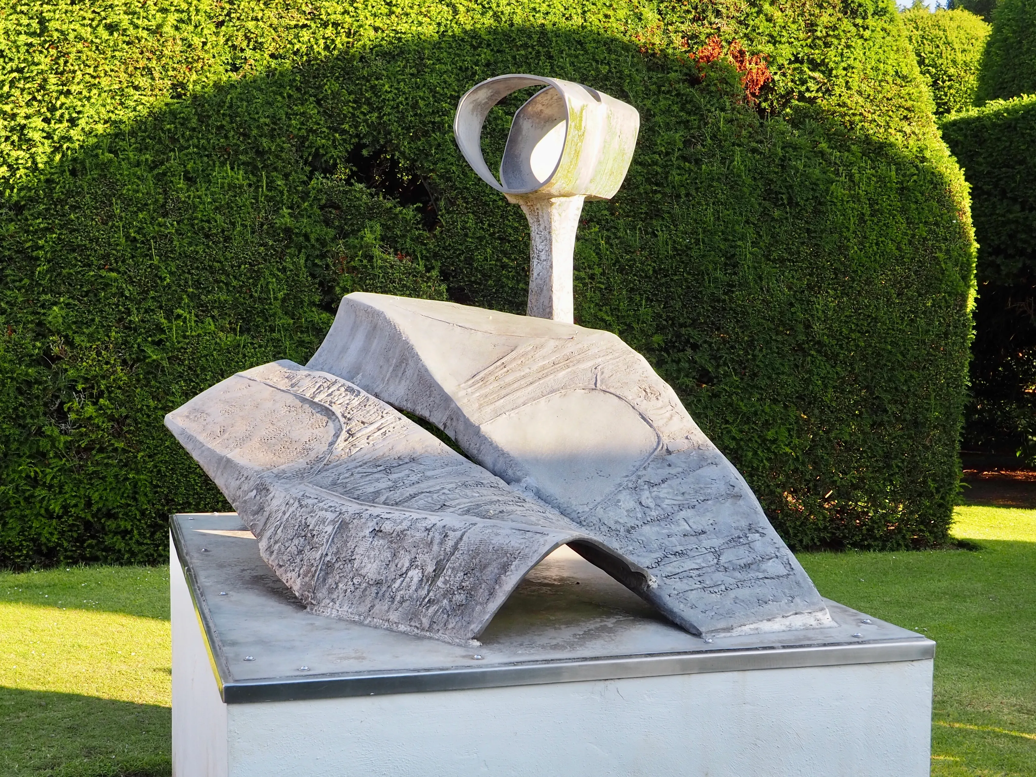 Escultura abstracta al aire libre con una base plegada de textura gris y un bucle de metal pulido que surge de ella, exhibida sobre un pedestal blanco frente a setos verdes perfectamente cuidados.