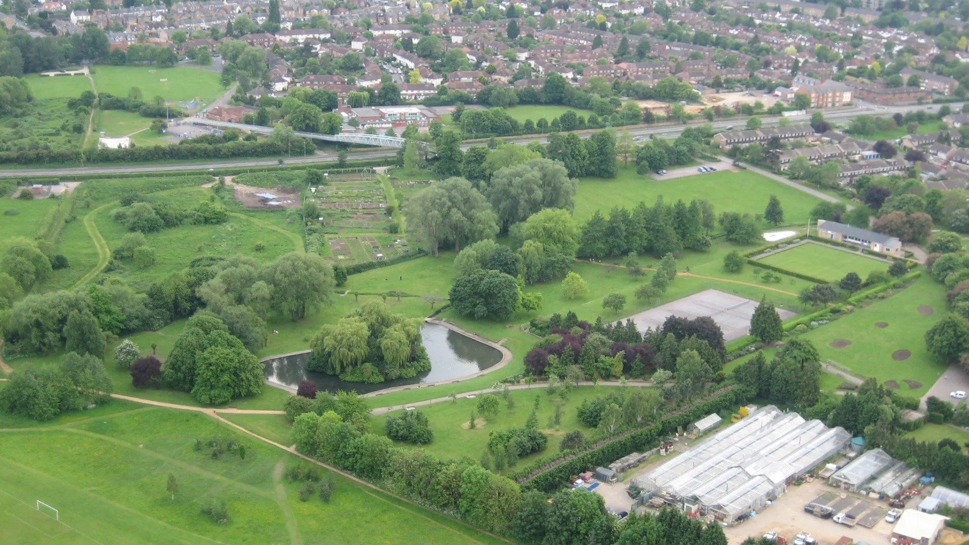 Luchtfoto van de Oxford University Parks met groene velden, een vijver, kassen en de omliggende woonwijken.