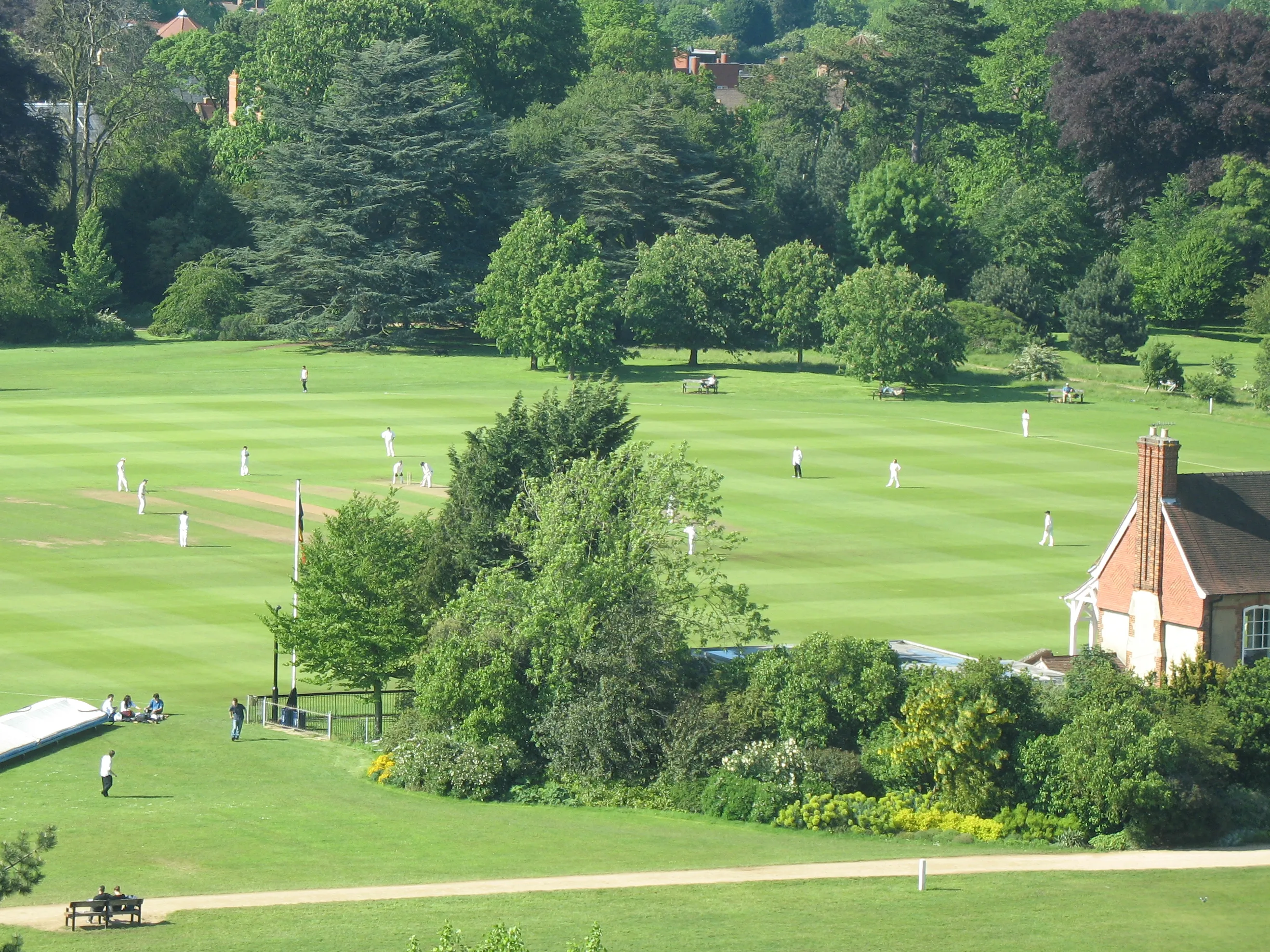 Cricketwedstrijd op een groot groen sportveld omringd door volwassen bomen, met een rood bakstenen gebouw aan de rechterkant.