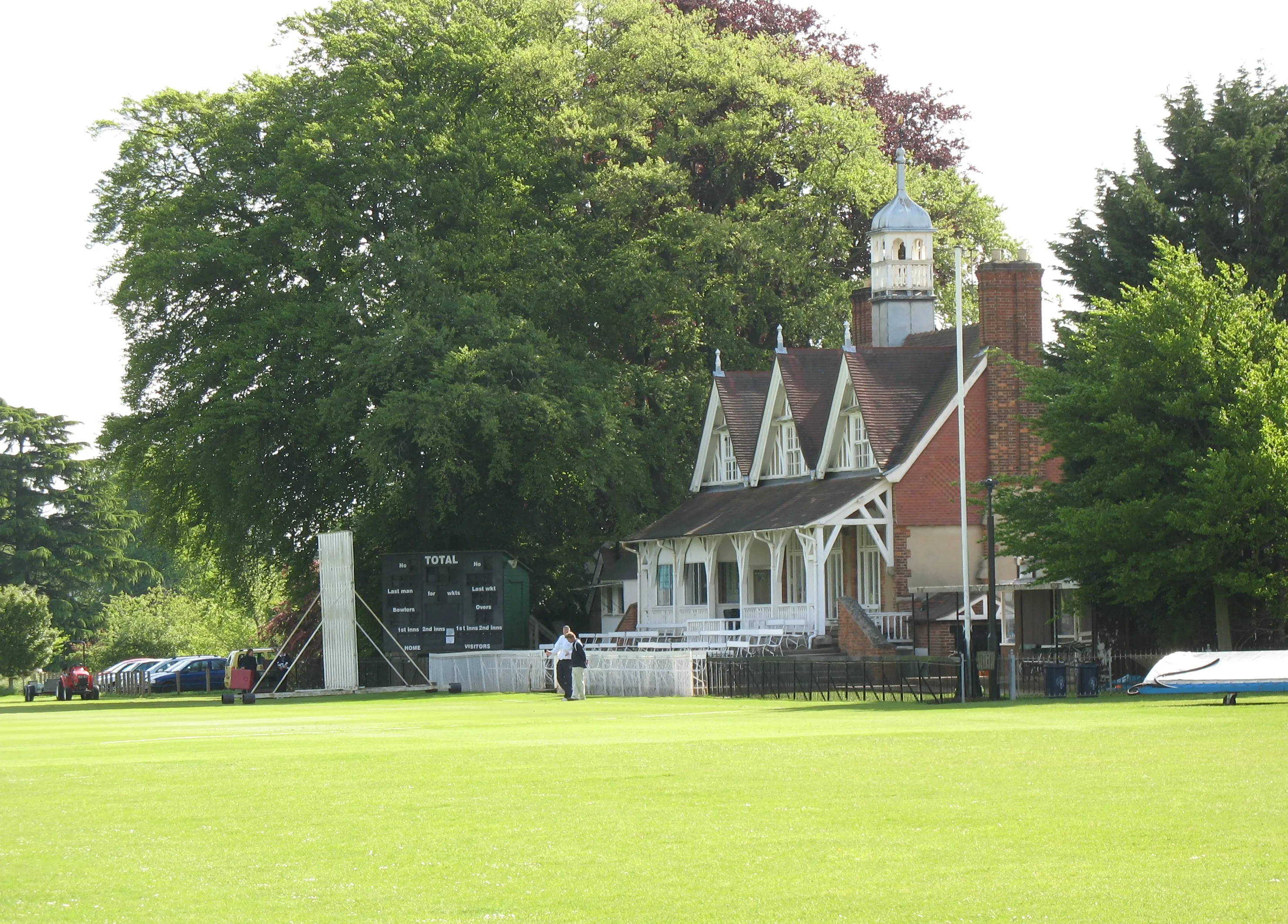 Bakstenen cricketpaviljoen met witte accenten en een koepeltoren, gelegen tussen grote bomen met een groen cricketveld op de voorgrond in de Oxford University Parks.