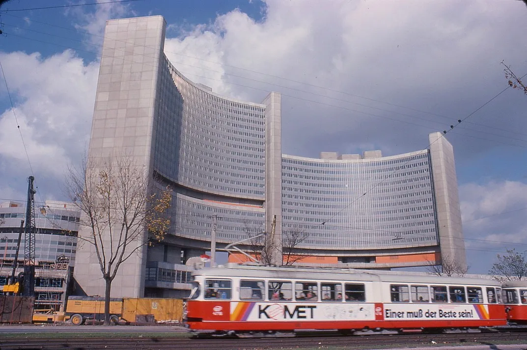 Een modern gebogen gebouw met een glazen gevel, een rode en witte tram op de voorgrond, en een blauwe lucht met wolken.