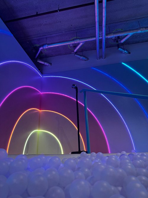 Colorful neon arches in purple, blue, orange, and yellow illuminate an indoor space with a white ball pit and exposed ceiling pipes.