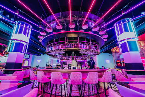 Vibrant interior space with pink and purple neon lighting, featuring a circular bar with upholstered stools and reflective surfaces.
