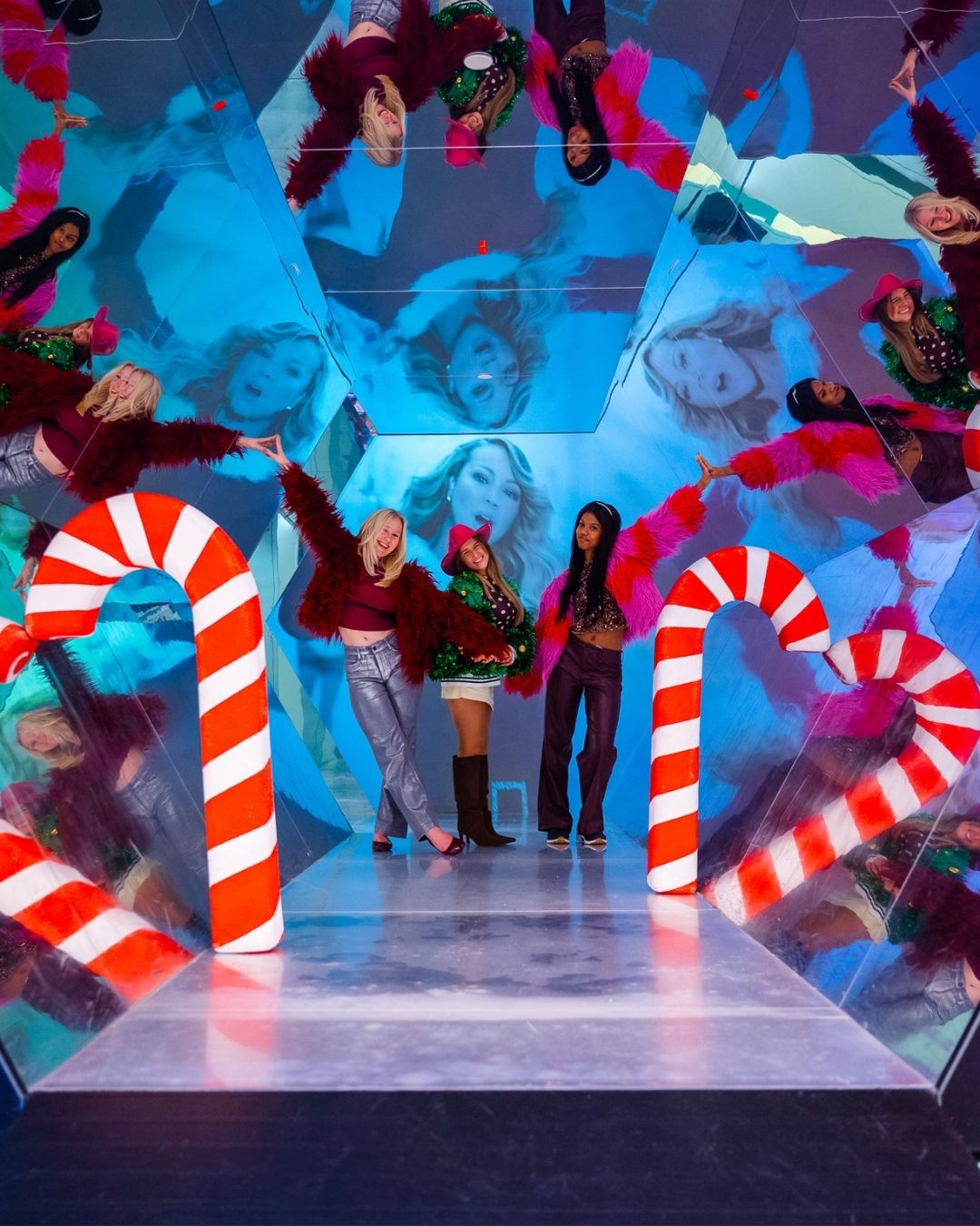 Three people posing among giant red-and-white candy canes in a mirrored room with colorful, distorted reflections and festive decor
