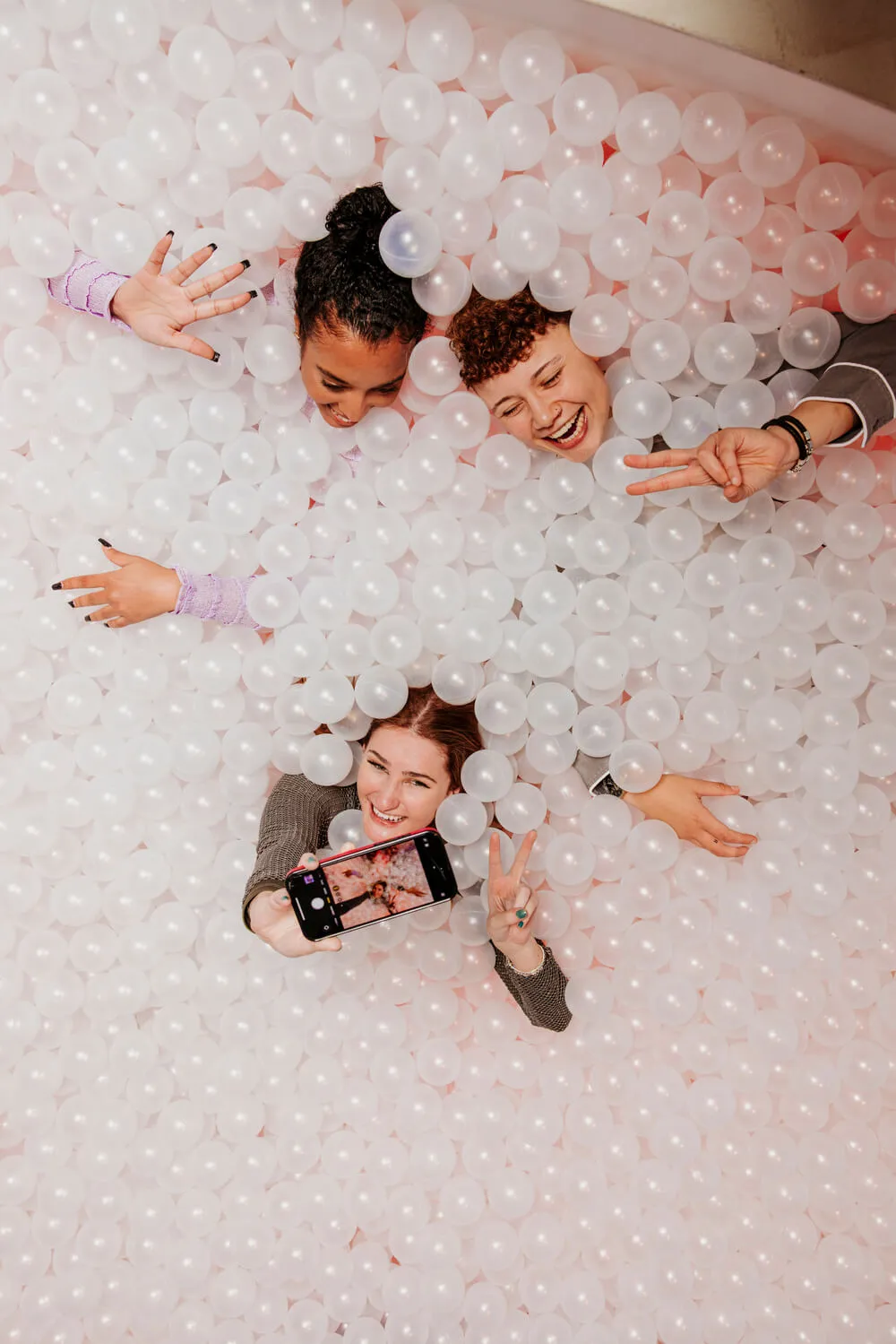 Three people interacting in a white and clear plastic ball pit; one holds a smartphone taking a selfie.