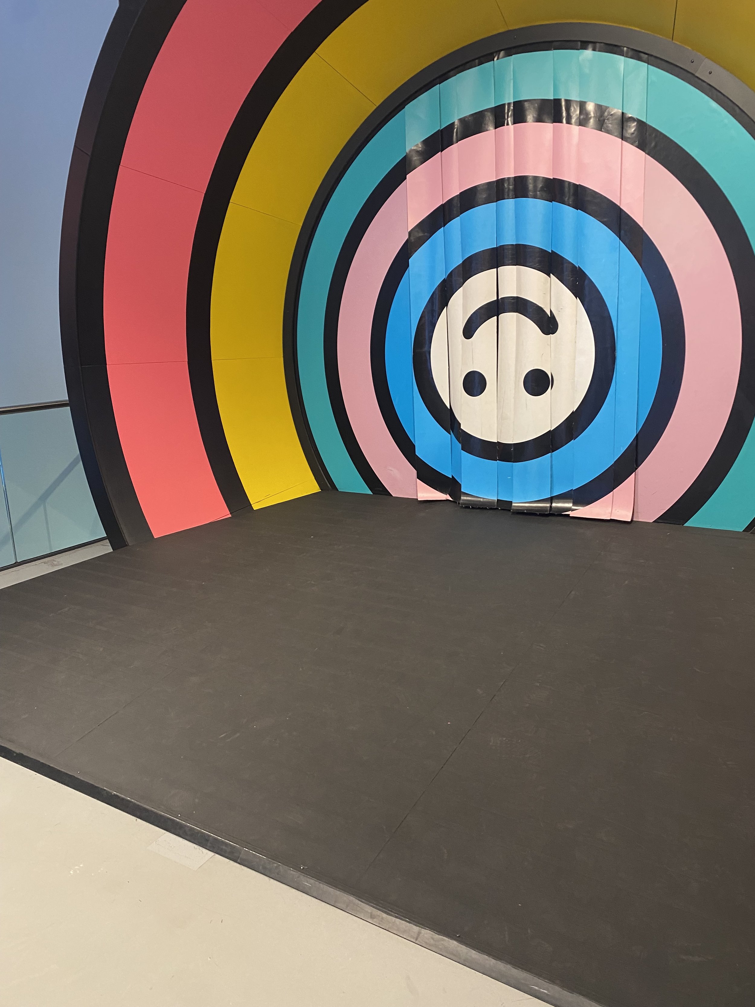 Colorful concentric circles in pink, yellow, turquoise, and blue form an optical illusion installation with a face-like center design in an indoor space