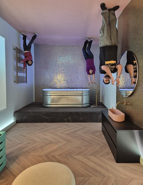 Upside-down bathroom scene with people appearing to hang from ceiling, featuring bathtub, sink, and herringbone wood flooring