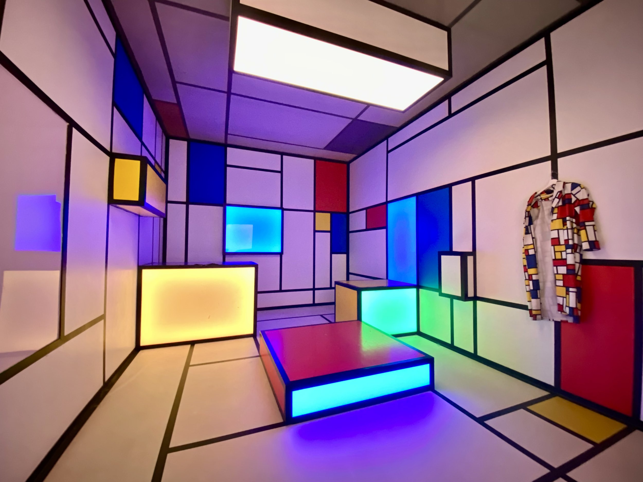 Colorful geometric room with blue, red, yellow, and green panels; checkered outfit hangs on the wall