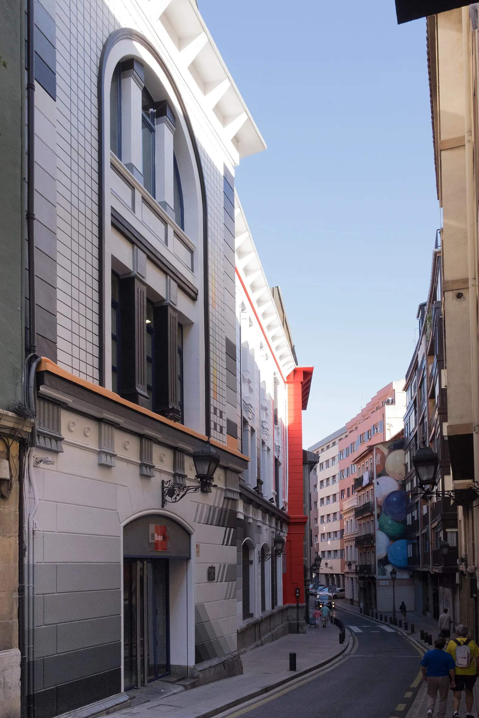 Vista callejera de un edificio con fachada pintada en trompe-l'œil que muestra ventanas arqueadas y detalles arquitectónicos, un elemento vertical rojo y coloridas esculturas de globos a lo largo de una estrecha calle urbana