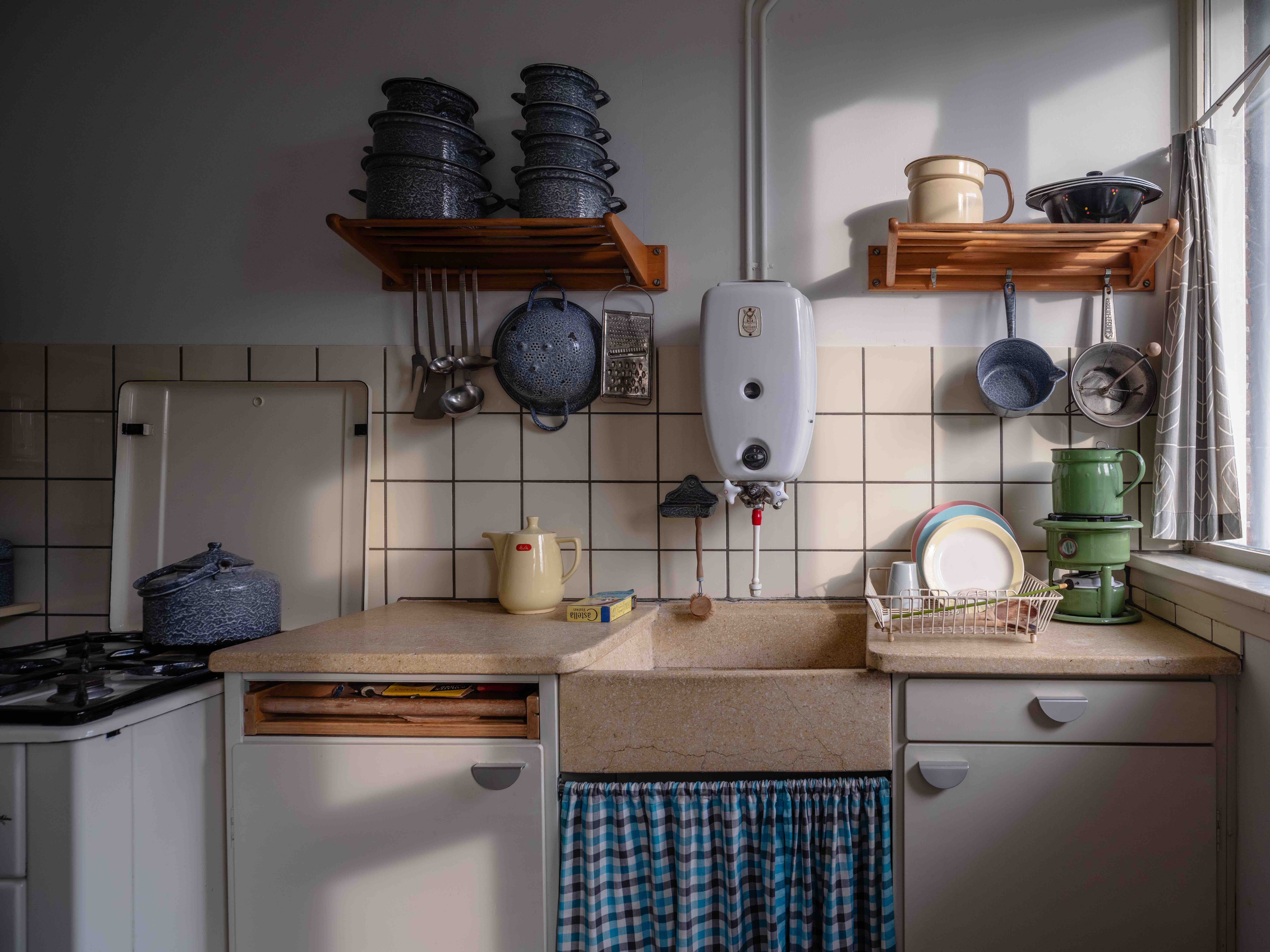 Exhibición de cocina vintage con paredes de azulejos blancos, estantes de madera con ollas, fregadero con cortina de cuadros azules y estufa antigua