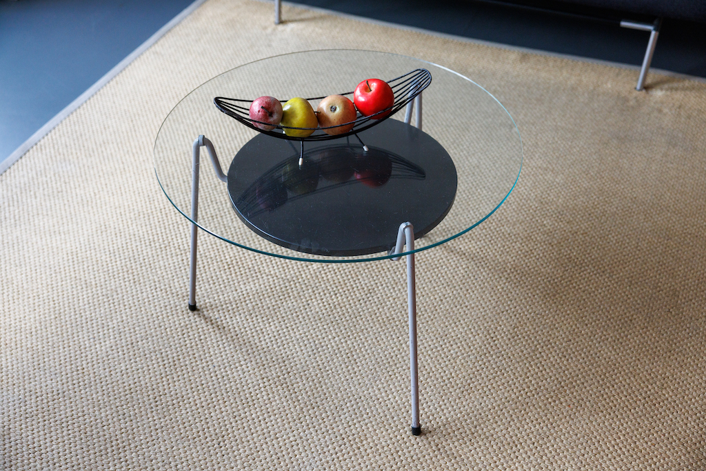 Mesa redonda de cristal con patas de metal, base circular negra y frutero de alambre con manzanas sobre alfombra beige