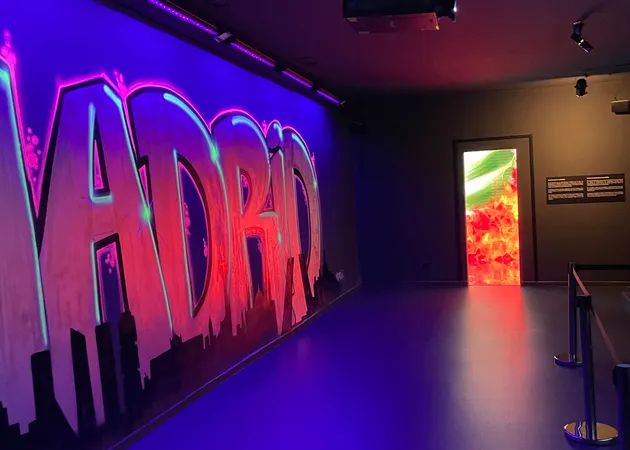 Neon-lit wall with the word 'MADRID' in large letters, illuminated in purple and pink, inside a modern museum gallery with a colorful projection visible through a doorway.