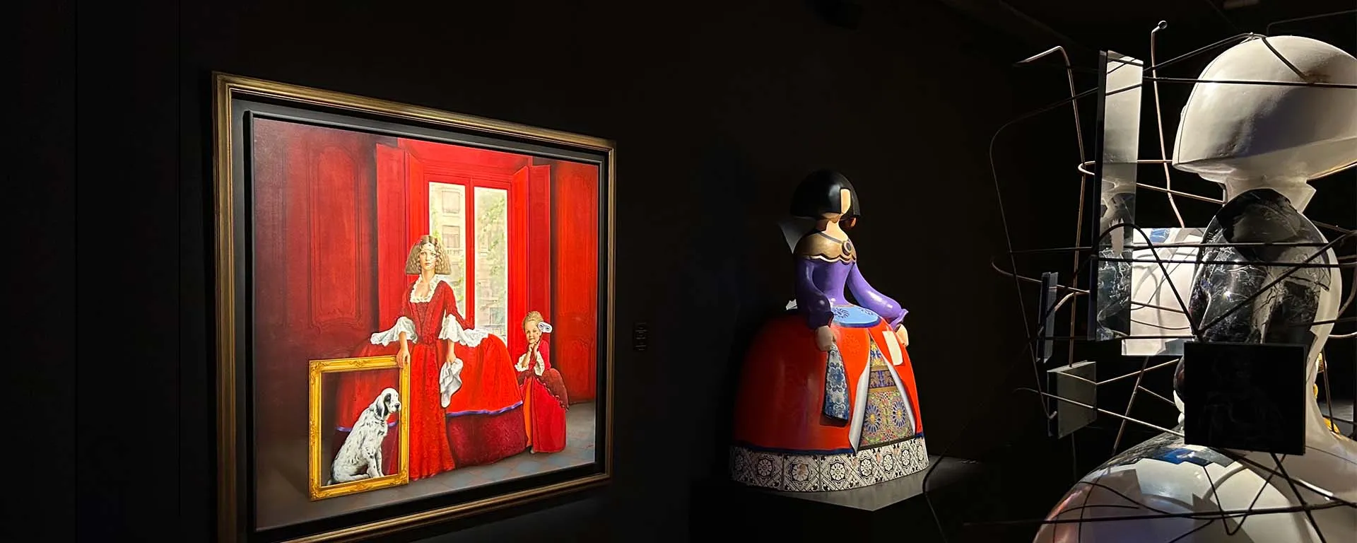 A framed painting of 'Las Meninas' displayed next to a sculptural figure and a wireframe structure in a dark gallery space