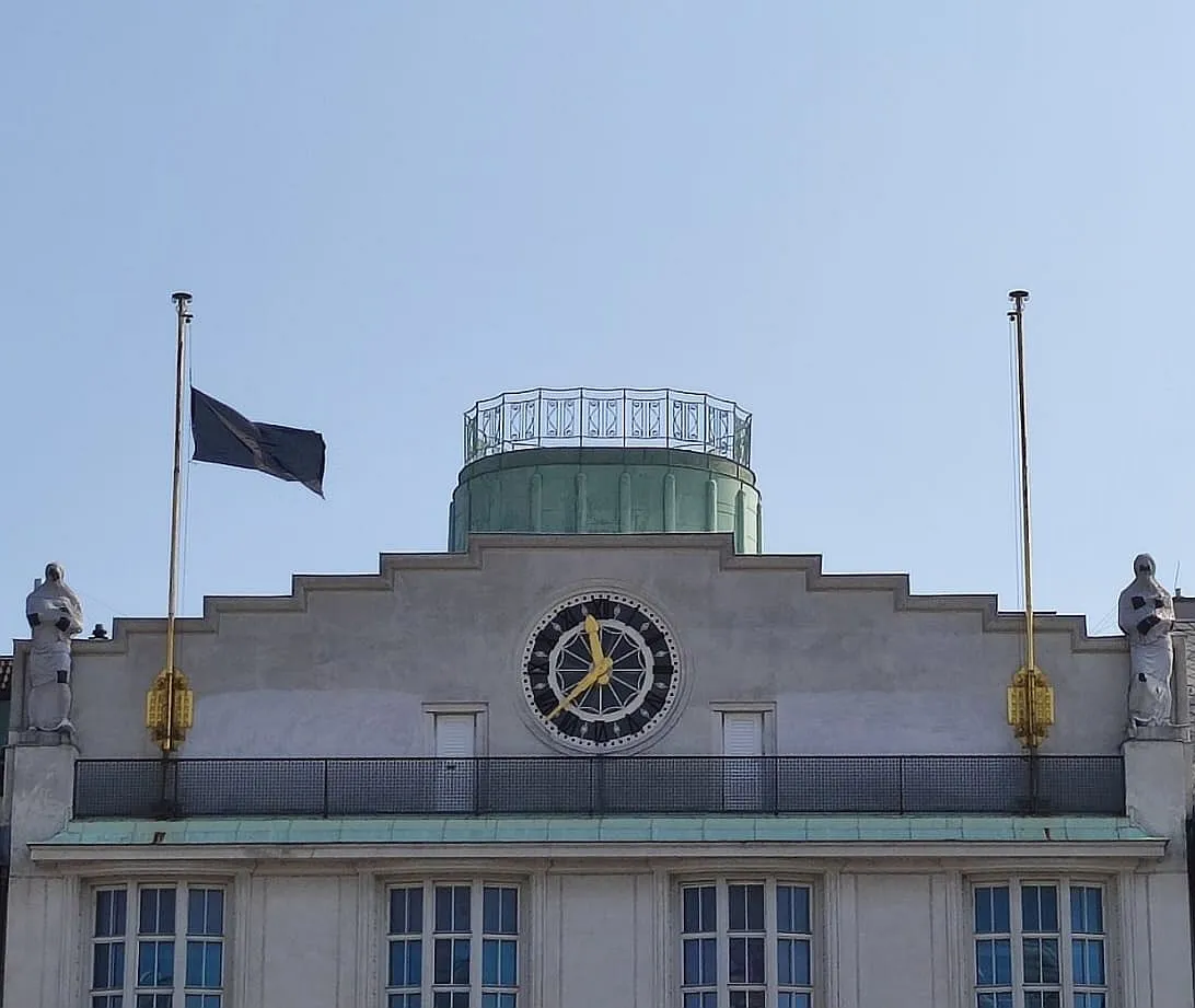 Fachada del edificio con un gran reloj, dos mástiles y estatuas bajo un cielo despejado