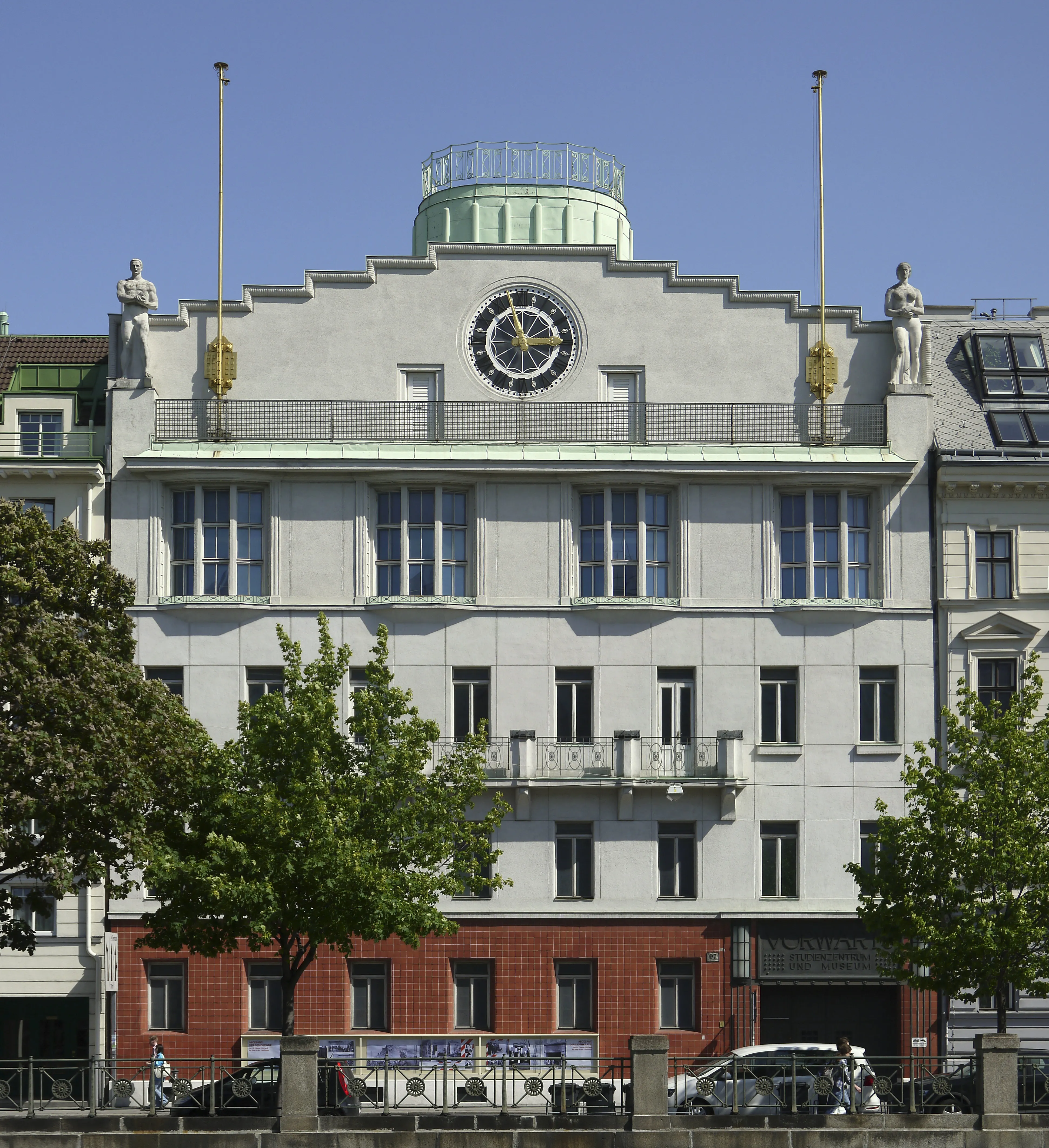 Un edificio blanco con un gran reloj y estatuas decorativas, con una cúpula verde en la parte superior, y árboles en primer plano.