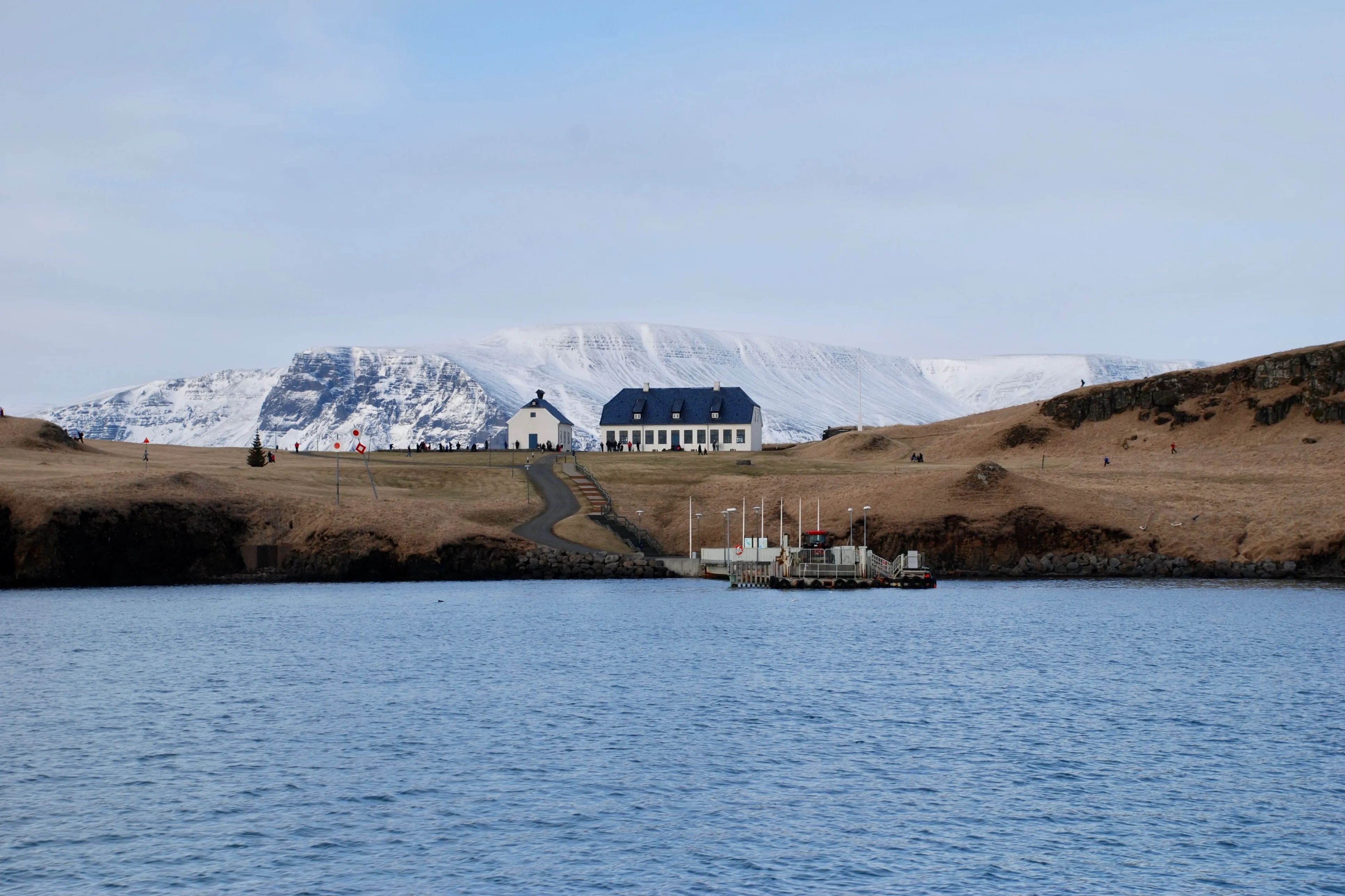 Vista de la isla de Viðey desde el agua, donde se aprecia una iglesia blanca, un edificio grande de tejado oscuro y un muelle de madera, con montañas nevadas al fondo.