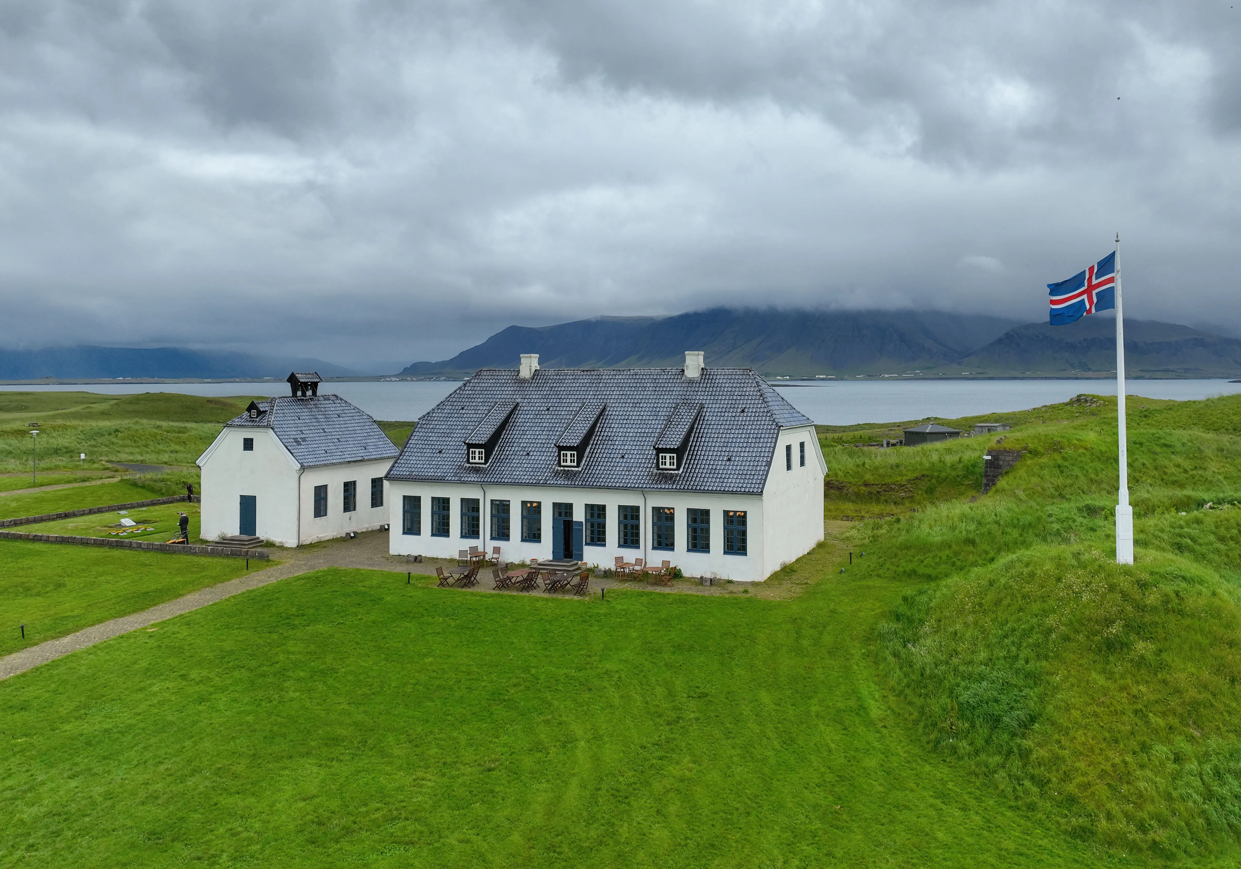 La histórica casa blanca Viðey House con un techo oscuro y una bandera islandesa ondeando en un mástil, situada sobre hierba verde con montañas y agua de fondo bajo un cielo nublado.