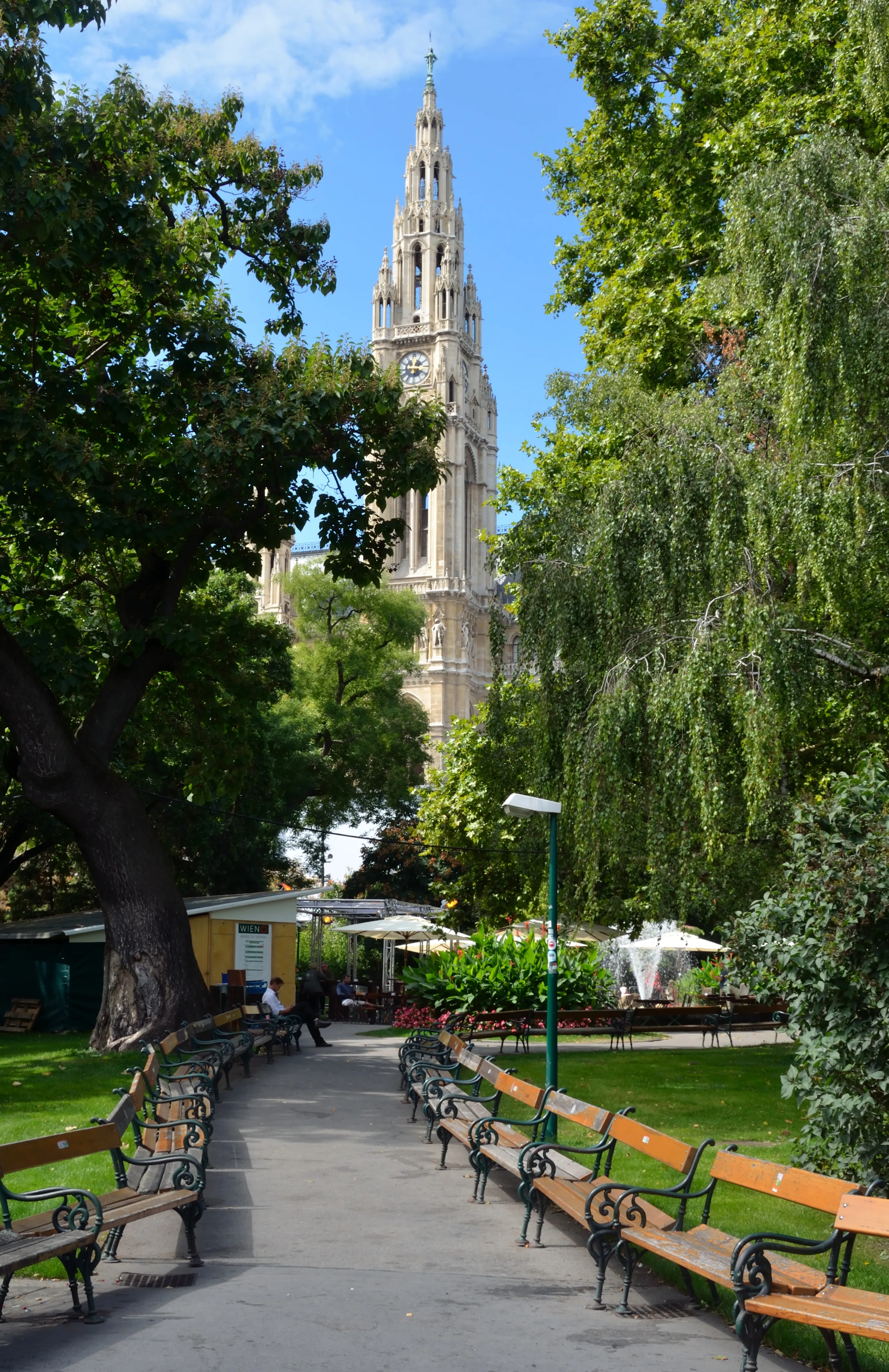A tall, ornate Gothic-style tower of Vienna City Hall seen through lush green trees and a park pathway with benches.