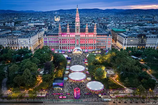 Aerial view of Vienna City Hall illuminated at dusk with pink and purple lights, surrounded by a festive market and crowds in the square.