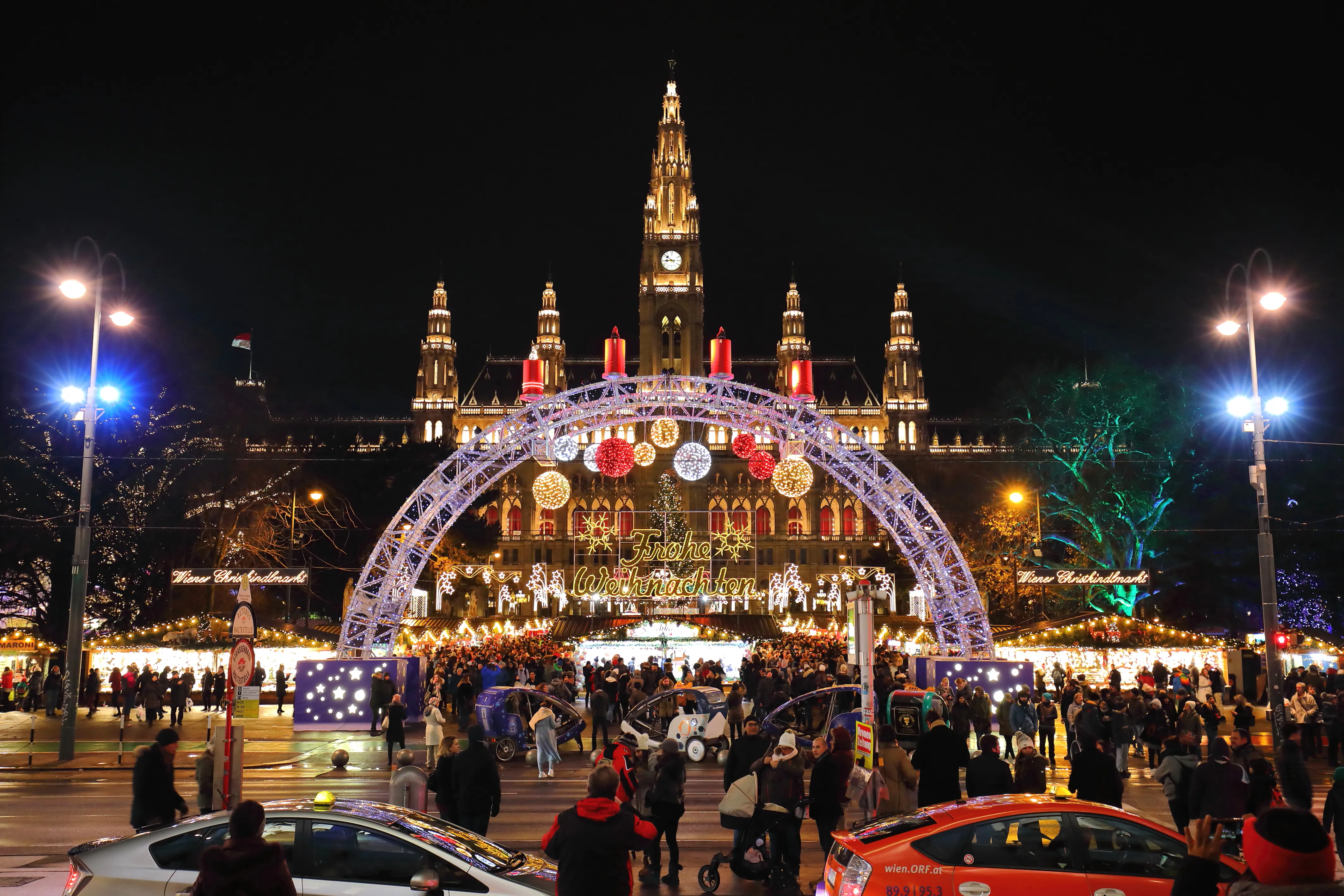 Vienna City Hall at night with a Christmas market, featuring a large decorated arch, lights, and people on Rathausplatz.