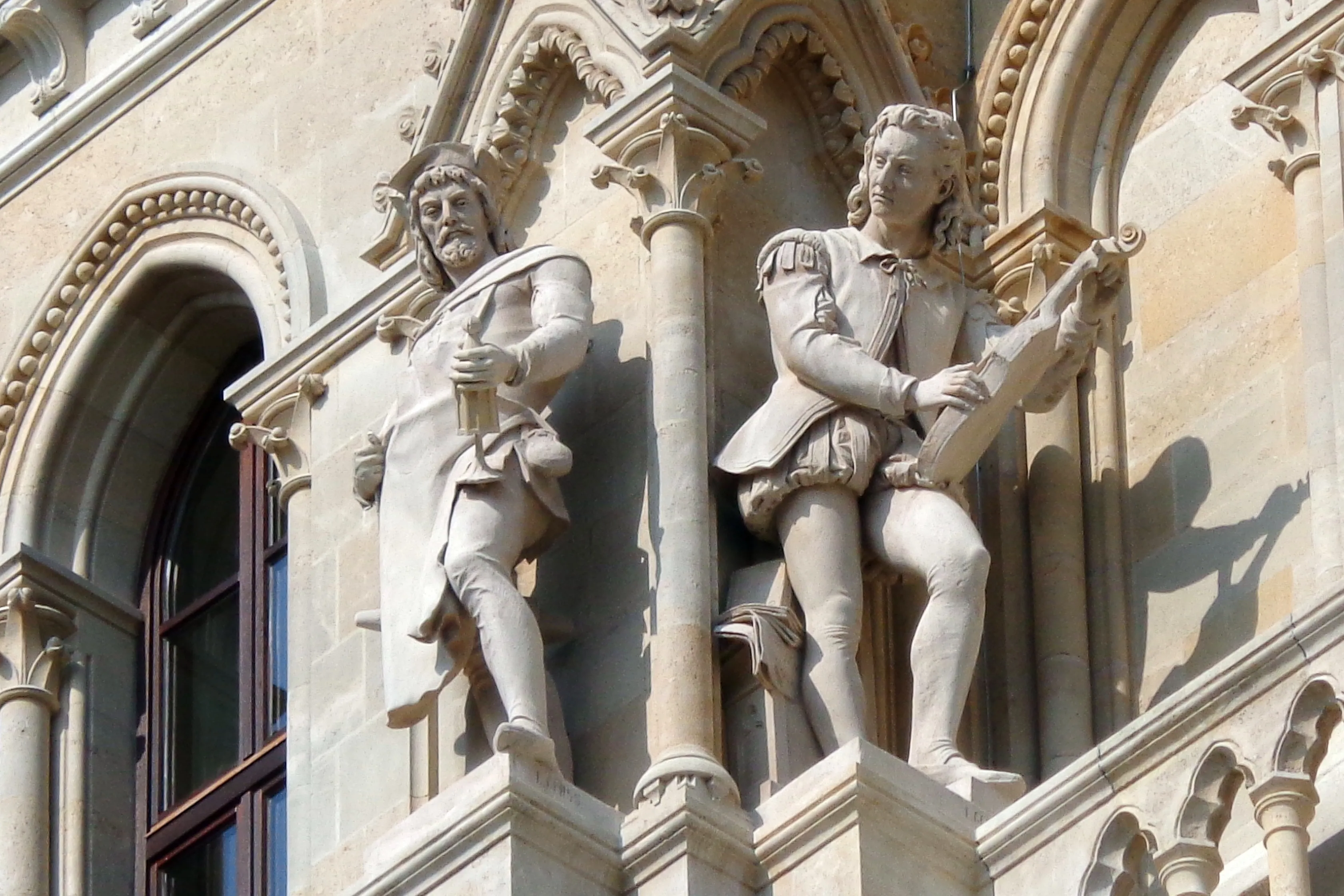 Two stone statues of historical figures on the facade of Vienna City Hall, with ornate architectural details and arched windows in the background.