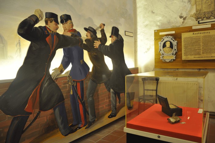 Una exhibición de museo que presenta un diorama de figuras históricas en uniformes policiales en una confrontación, junto a una vitrina con artefactos y un retrato enmarcado.