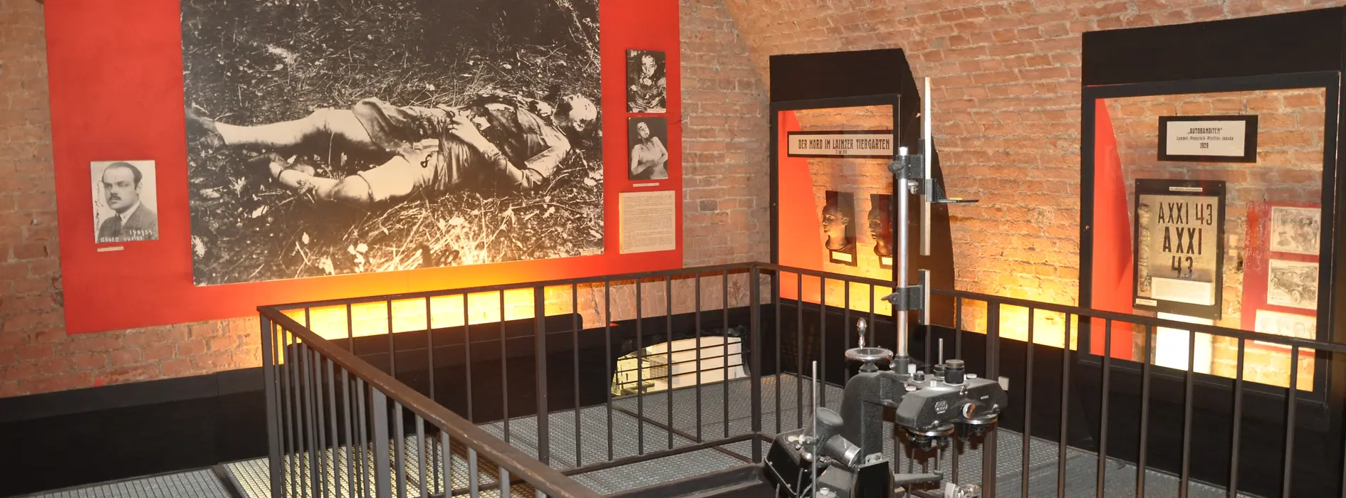 Interior de un museo con paredes de ladrillo, una gran fotografía en blanco y negro sobre una pared roja, vitrinas y una barandilla de metal