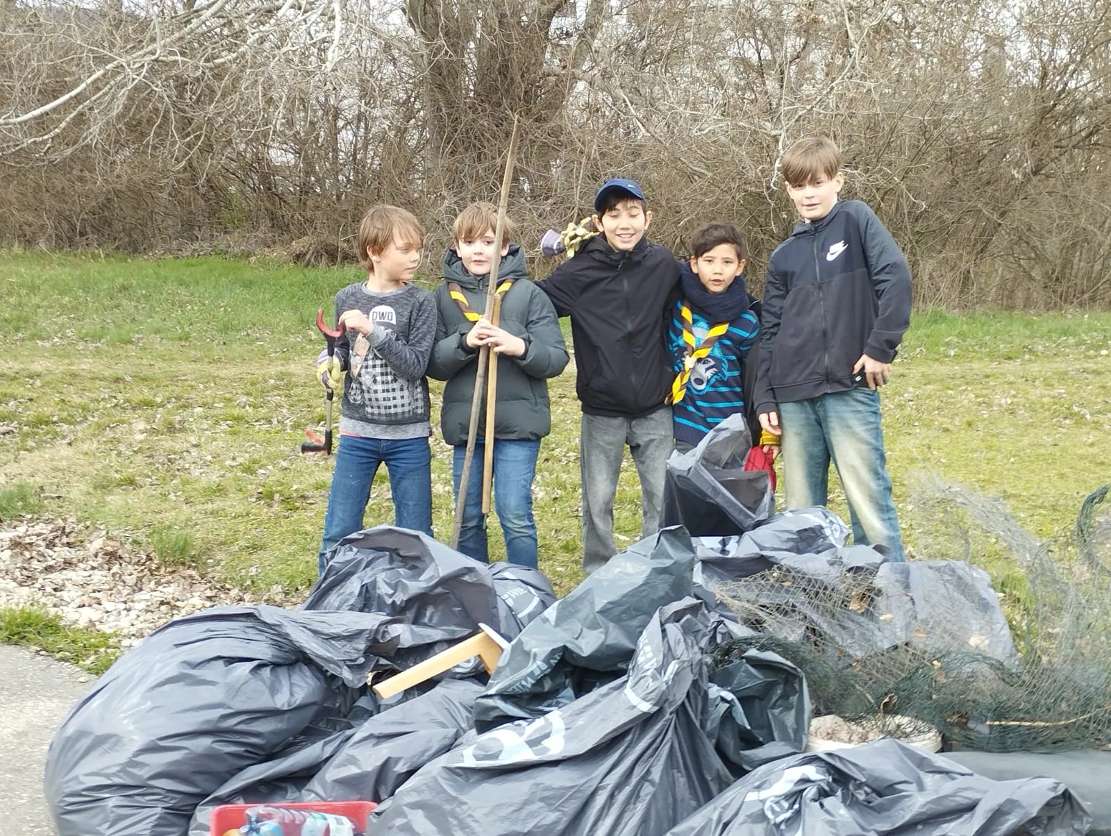 cinco niños parados al aire libre con bolsas de basura y herramientas de limpieza