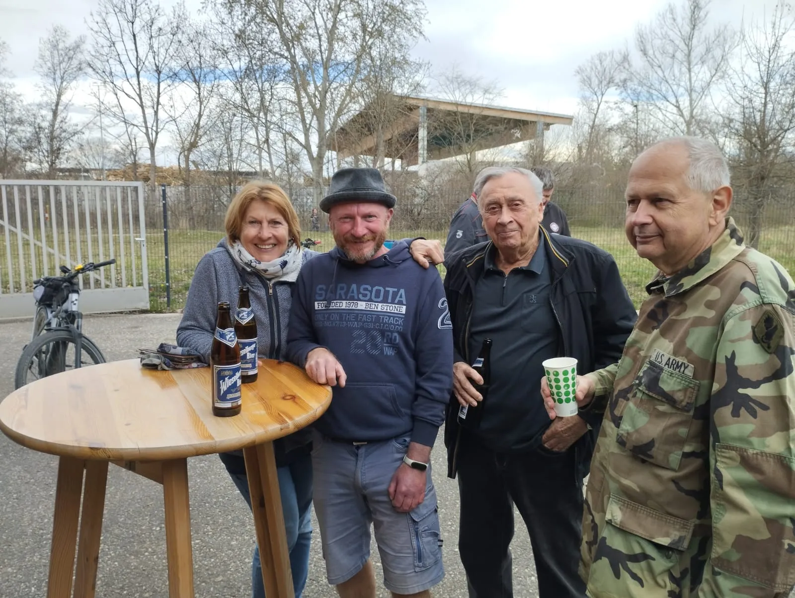 cuatro personas de pie al aire libre cerca de una mesa de madera con botellas de cerveza