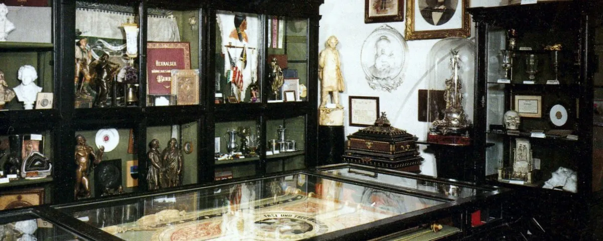 Interior de un museo con vitrinas de cristal que contienen trofeos, esculturas y artefactos históricos, incluida una gran mesa central con un mapa o documento.