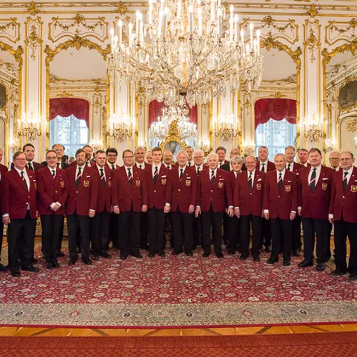 Miembros del coro con chaquetas de color granate de pie en un gran salón con candelabro y decoraciones ornamentadas