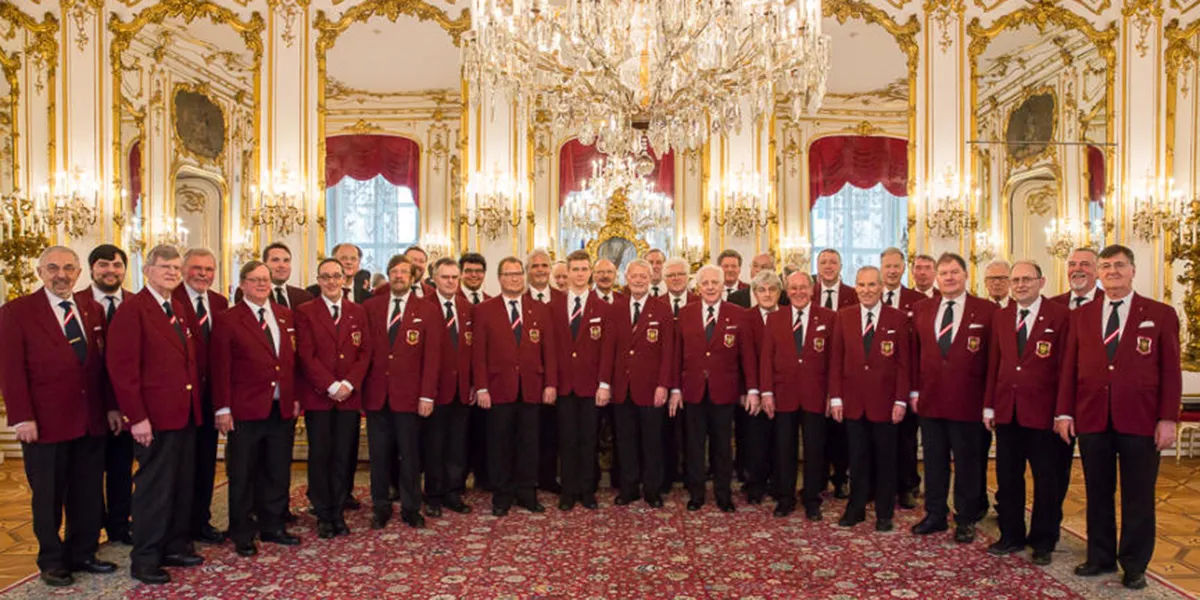 Grupo de hombres con chaquetas granates de pie en un gran salón con decoraciones doradas y una lámpara de araña
