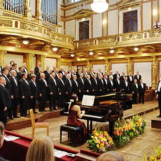 Un gran coro masculino con esmóquines negros actuando en un escenario con un piano de cola, elaboradas decoraciones doradas y arreglos florales.
