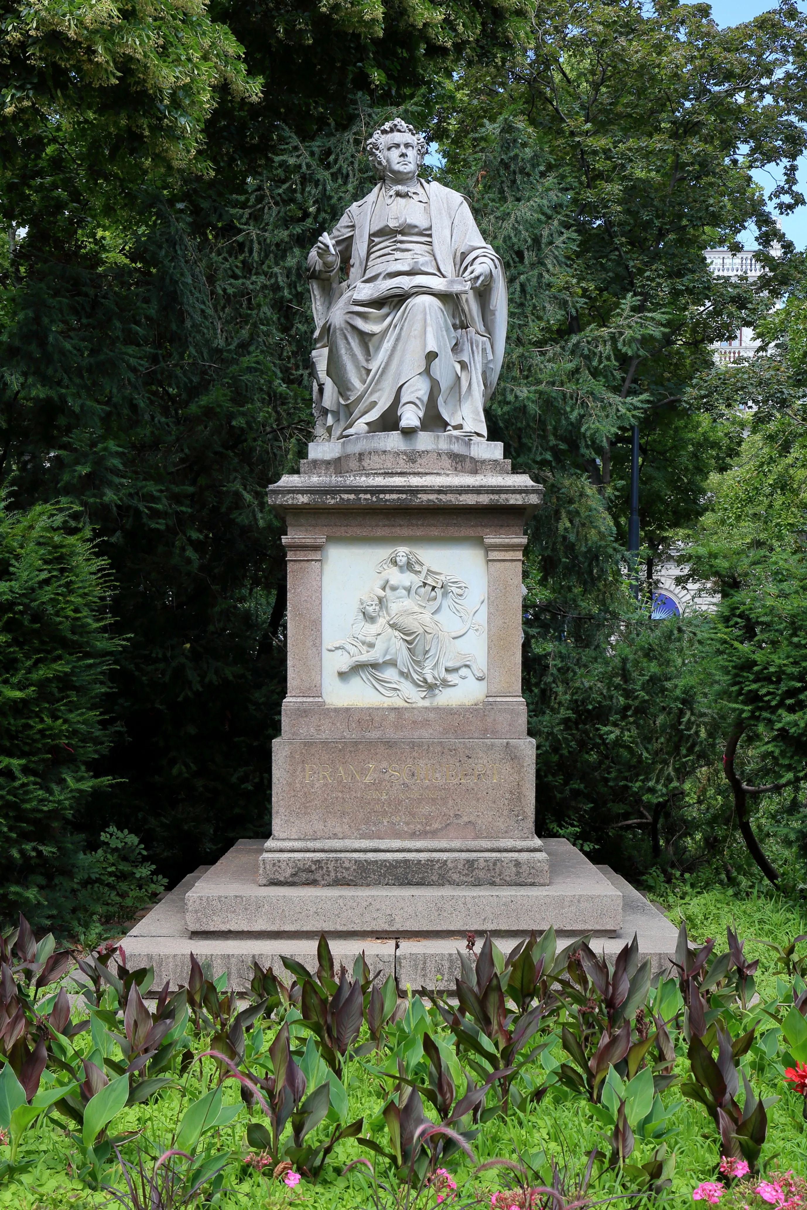 Una estatua de piedra de un hombre sentado sobre un pedestal con un relieve, rodeado de árboles y plantas verdes en un parque.