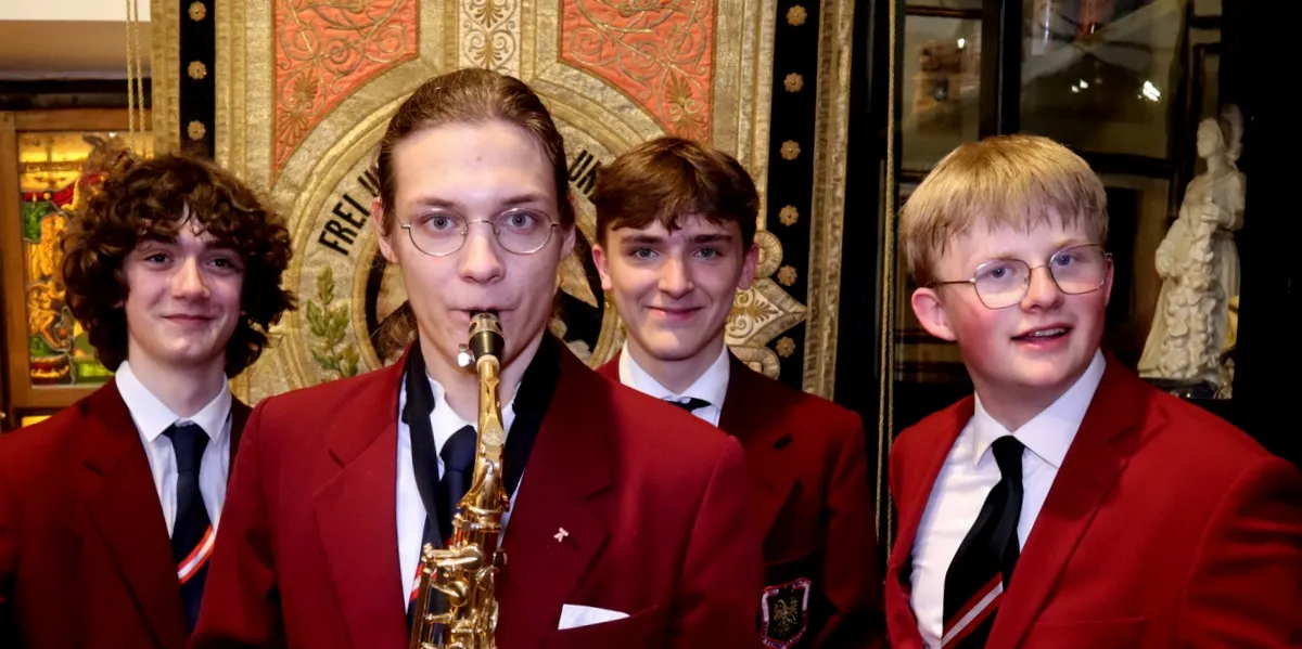 Vier jonge mannen in rode blazers staan binnen met een saxofoon en een sierlijke achtergrond