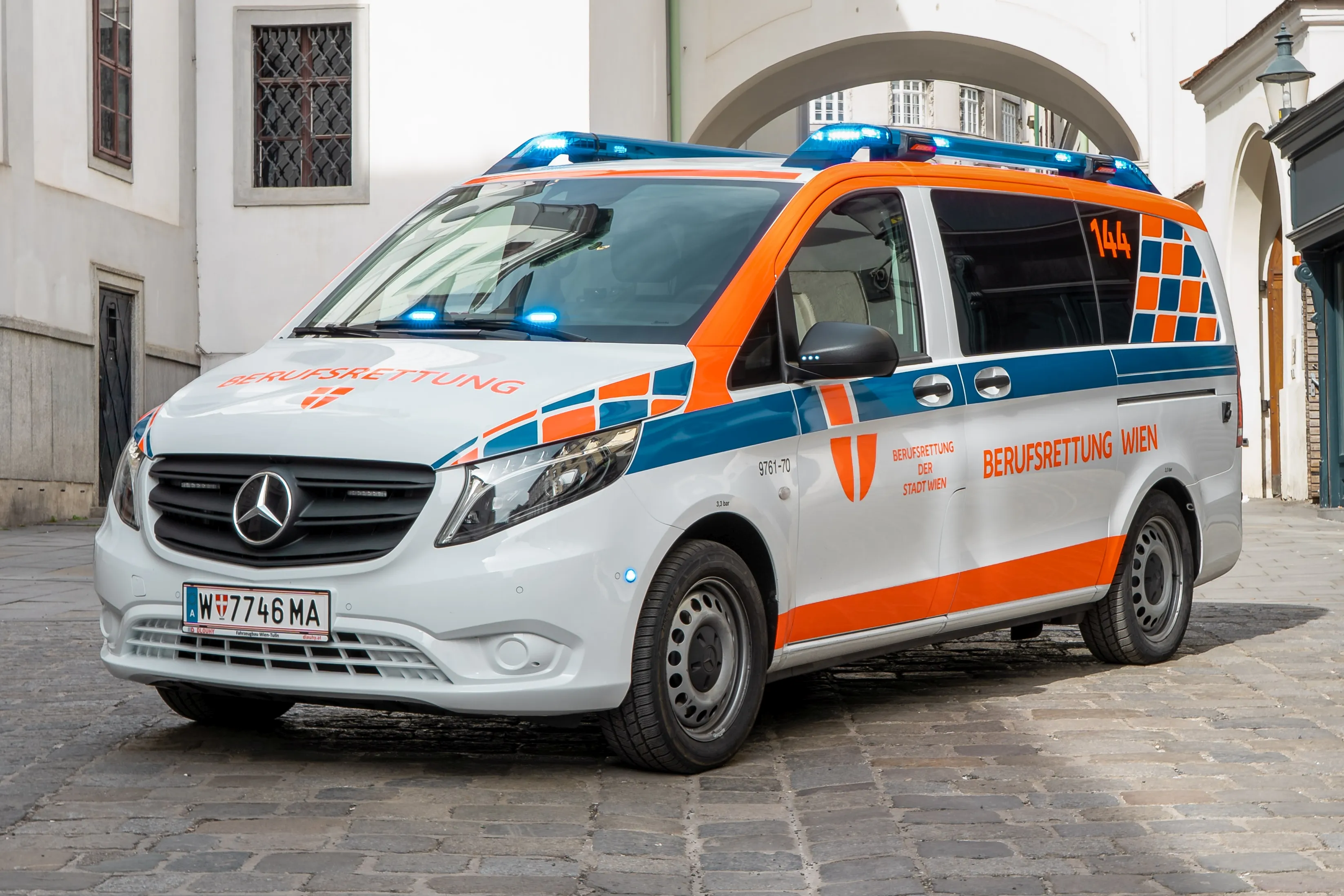 Una furgoneta blanca de emergencias con rayas naranjas y azules y el texto 'BERUFSRETTUNG WIEN' aparcada en una calle adoquinada.