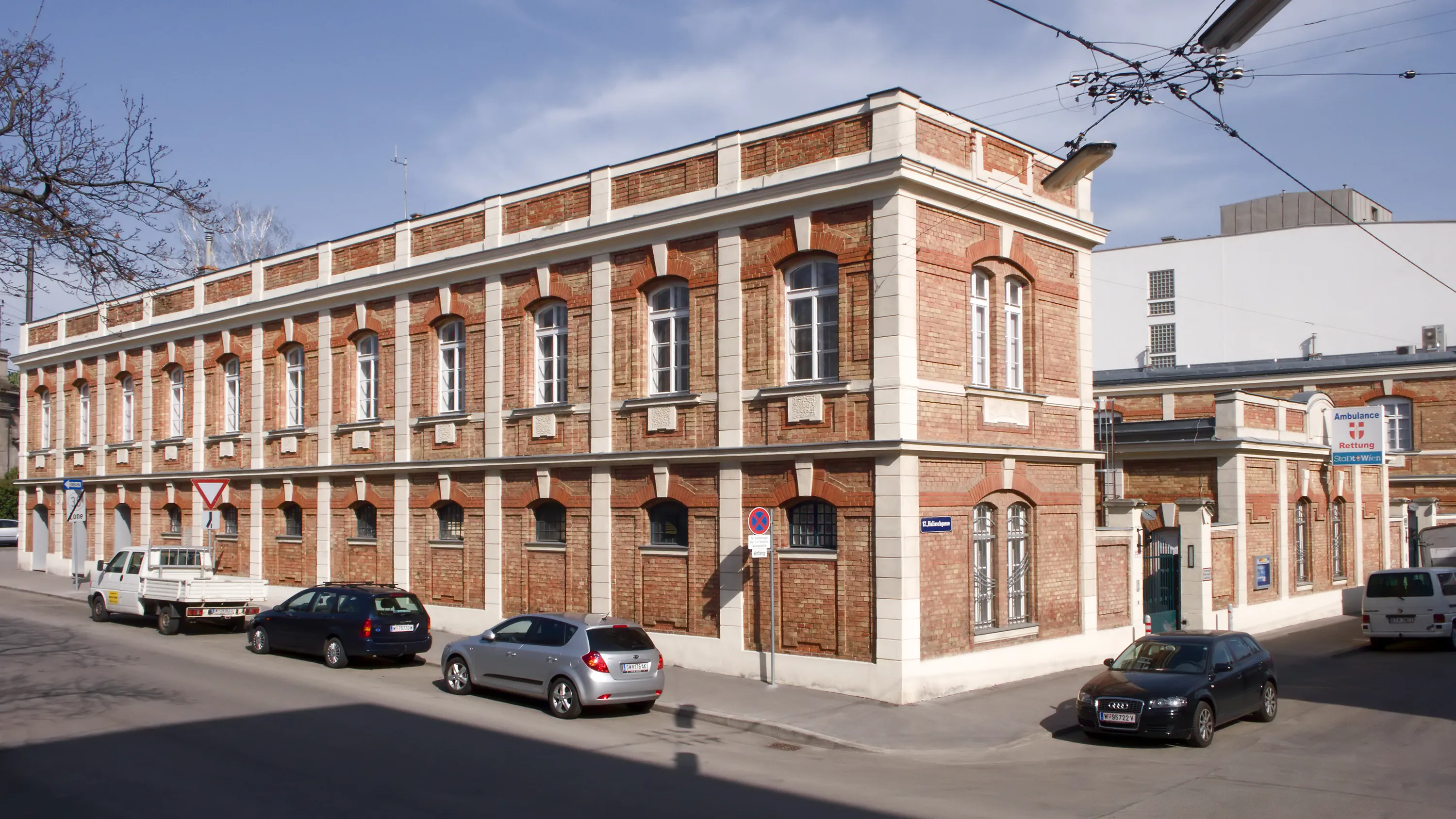 un edificio de ladrillo rojo con molduras blancas y ventanas arqueadas, con coches aparcados en la calle de enfrente
