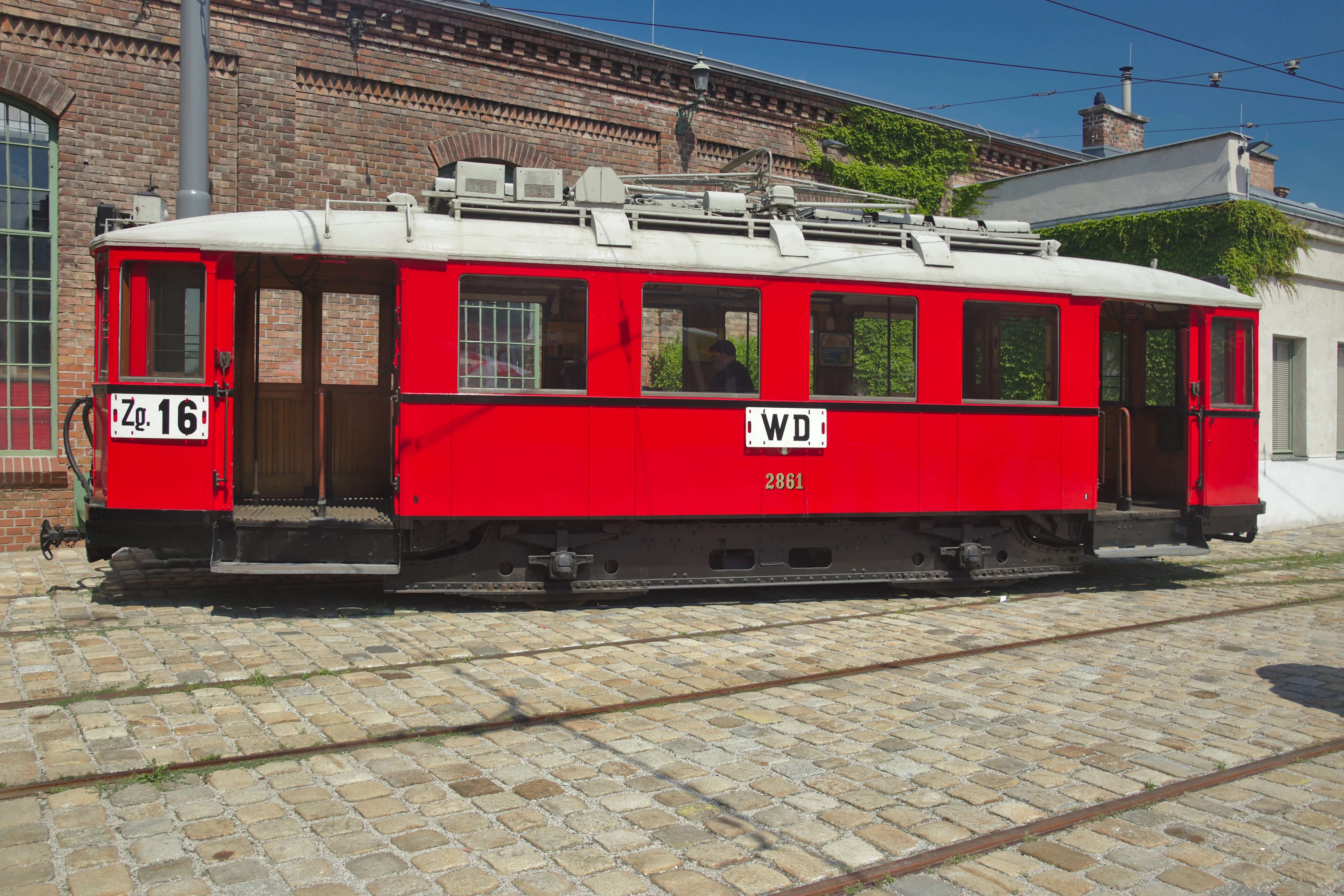 Czerwony zabytkowy tramwaj na torach przed ceglanym budynkiem