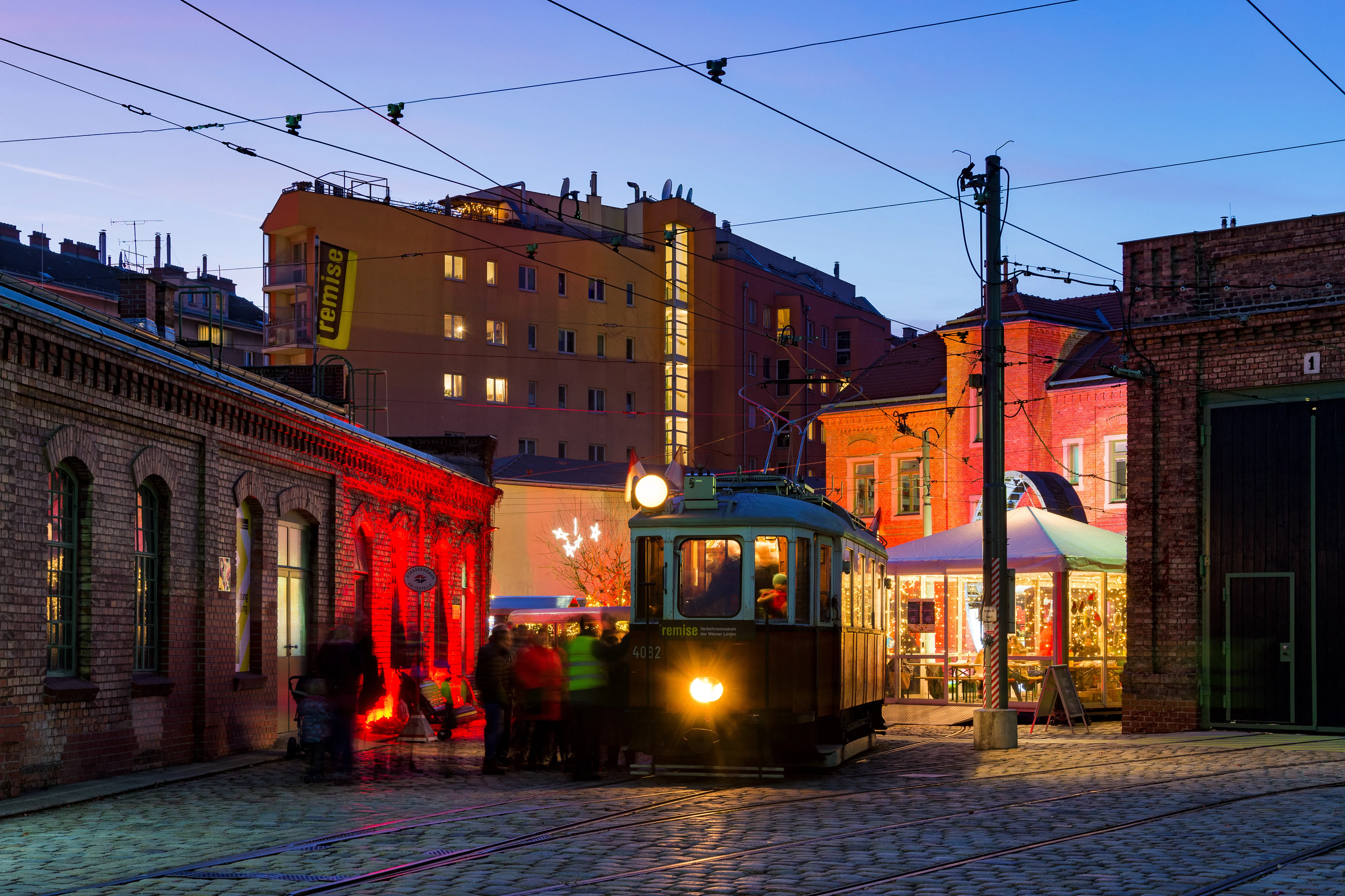 Zabytkowy tramwaj z włączonymi światłami zaparkowany jest na bruku przed ceglanym budynkiem o zmierzchu, z przewodami napowietrznymi i oświetlonym budynkiem w tle.