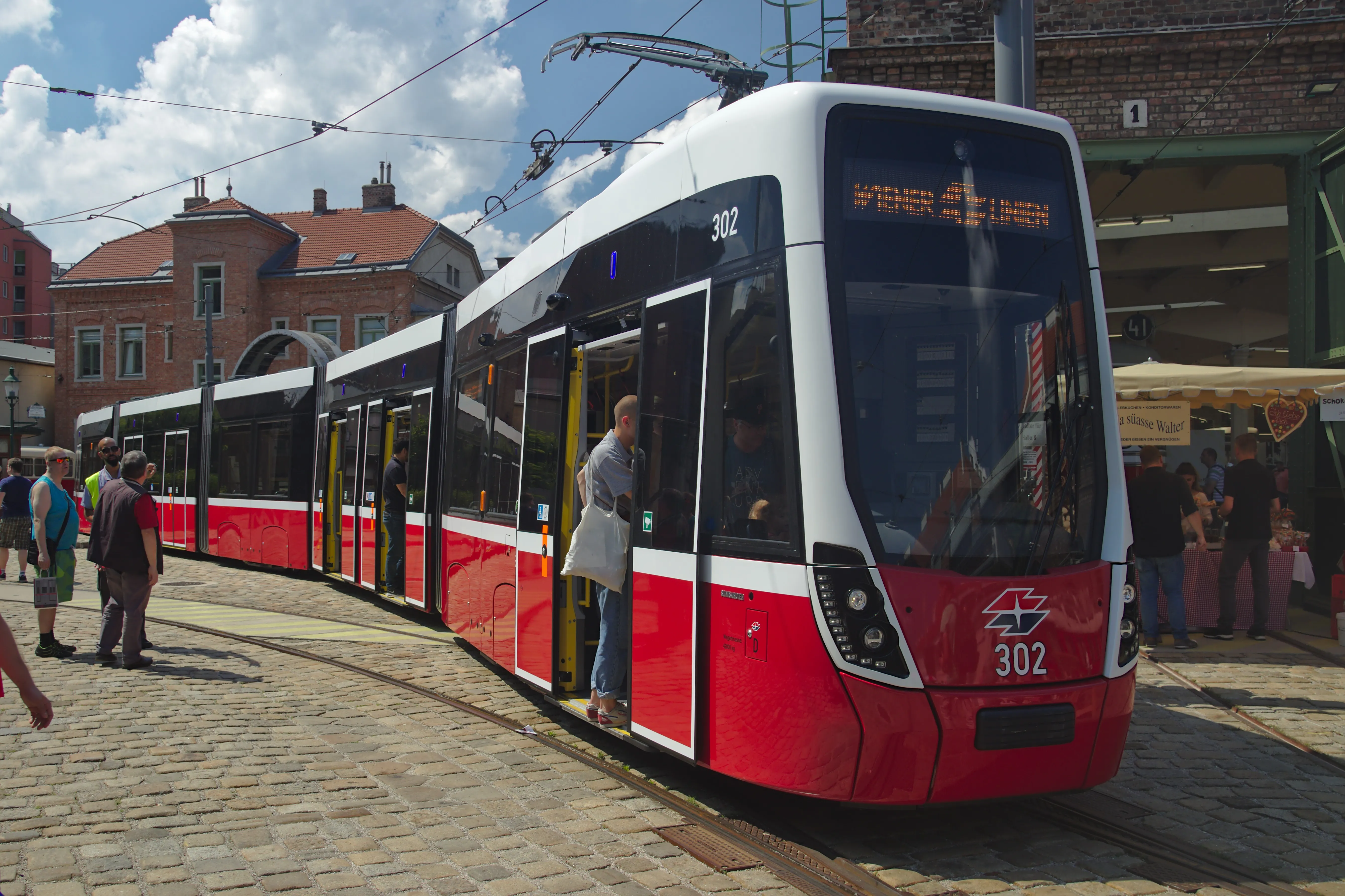 Een rode en witte moderne tram tentoongesteld in het Wiener Tramwaymuseum Remise, met mensen die in de buurt op een geplaveide straat staan.