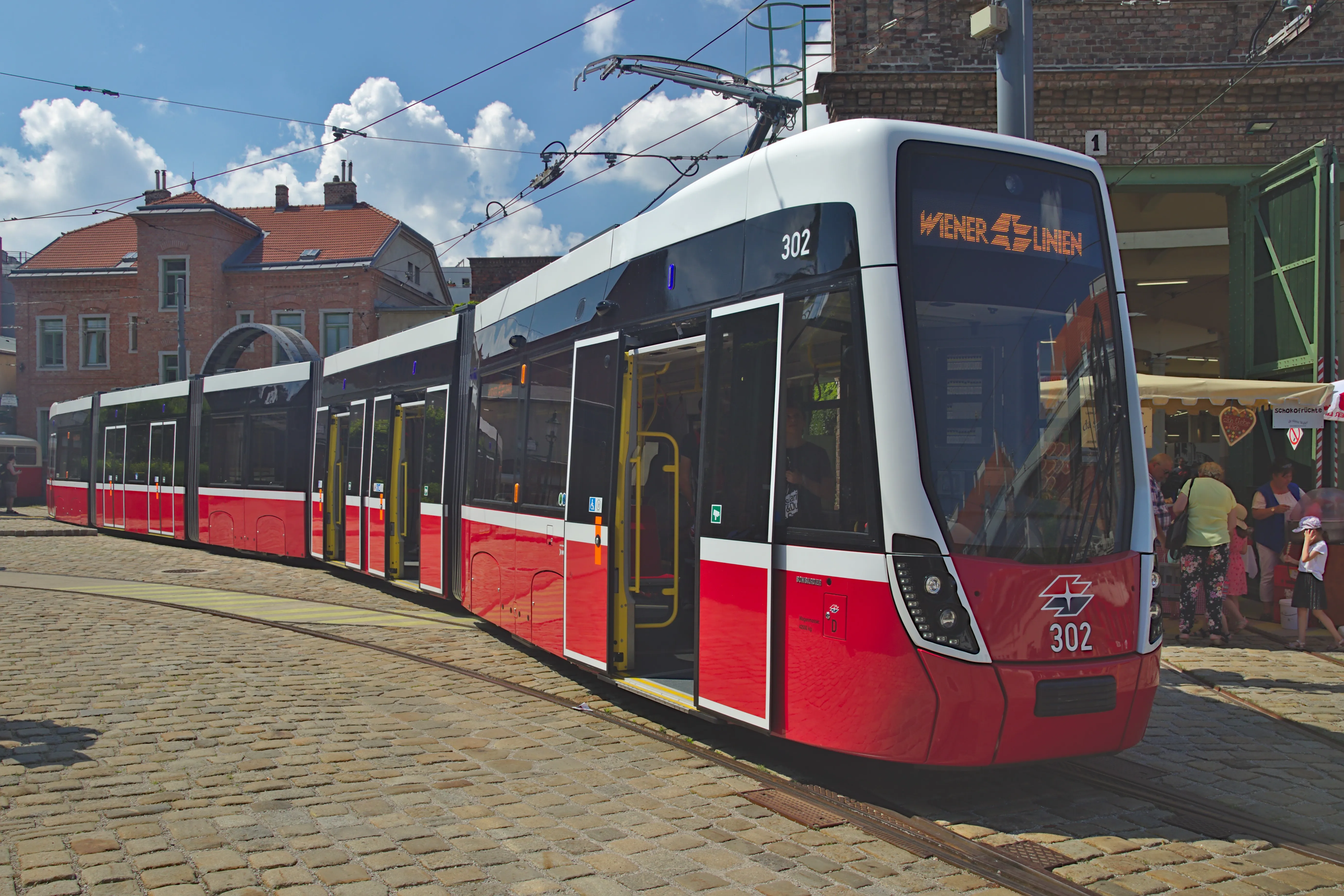 Een moderne rode en witte tram met nummer 302 staat op een geplaveide straat voor een bakstenen gebouw.