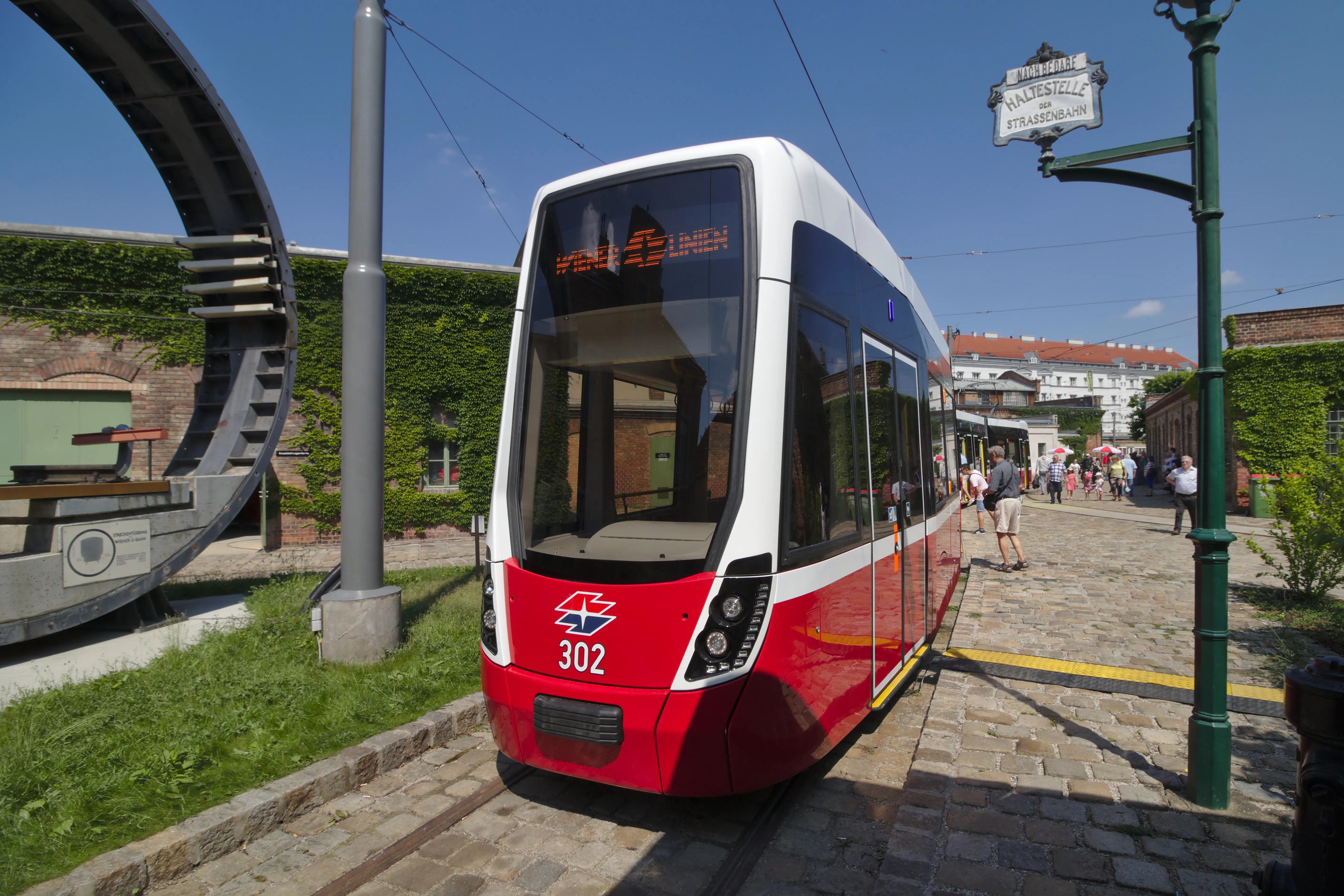 Czerwono-biały tramwaj numer 302 na wystawie przed budynkiem z zielonymi ścianami i dużą okrągłą rzeźbą