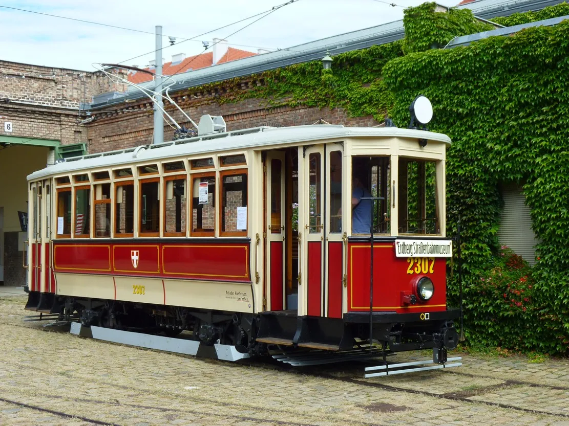 Czerwono-kremowy zabytkowy tramwaj z numerem 2307 stoi zaparkowany na torach przed ceglanym budynkiem z porośniętymi bluszczem ścianami.