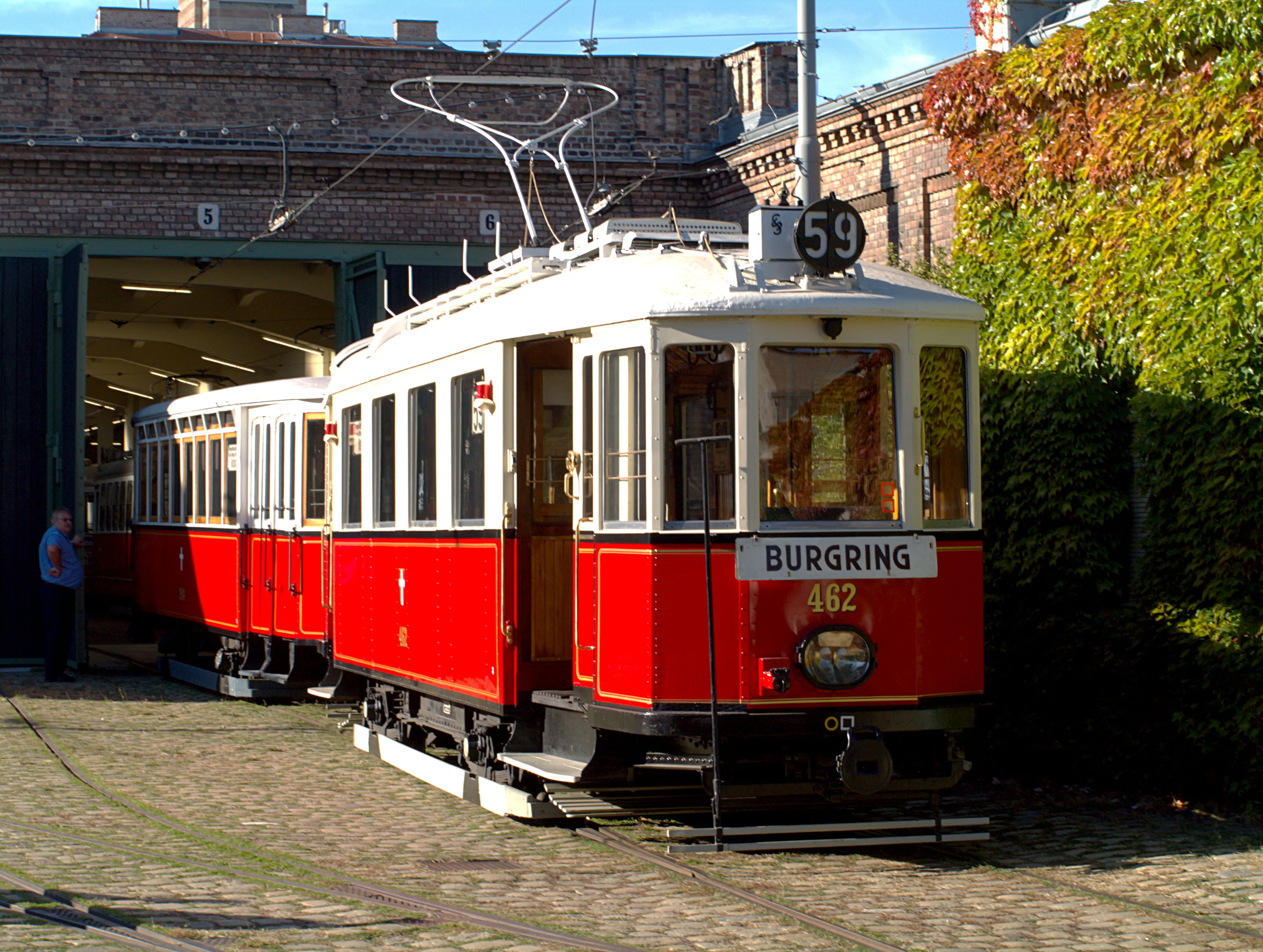 Czerwony i biały zabytkowy tramwaj z napisem 'BURGRING' i '462' zaparkowany przed ceglanym budynkiem z dużymi otwartymi drzwiami garażowymi.